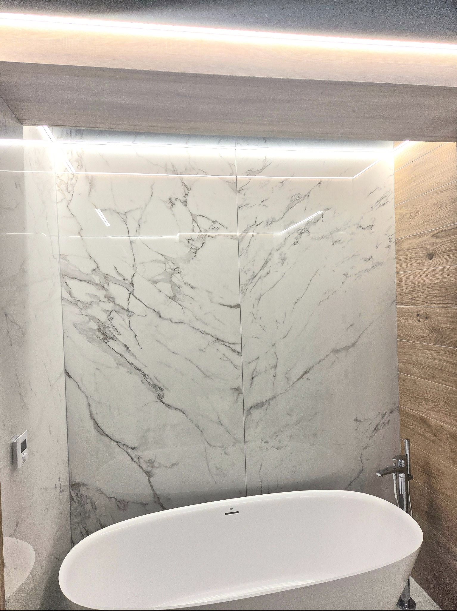 Nowoczesna łazienka z wanną wolnostojącą, marmurowymi płytkami na ścianach i oświetleniem LED. Minimalistyczny design, jasne kolory, drewniane akcenty.