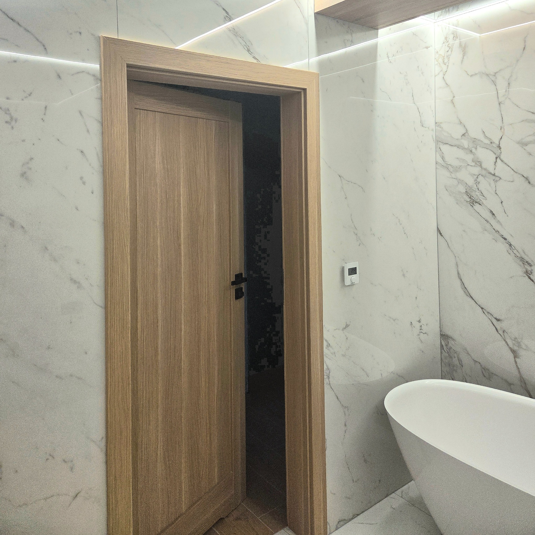 Nowoczesna łazienka z marmurowymi płytkami i drewnianymi drzwiami. Widoczna wanna wolnostojąca i subtelne oświetlenie LED. Minimalistyczny design z dbałością o detale.
