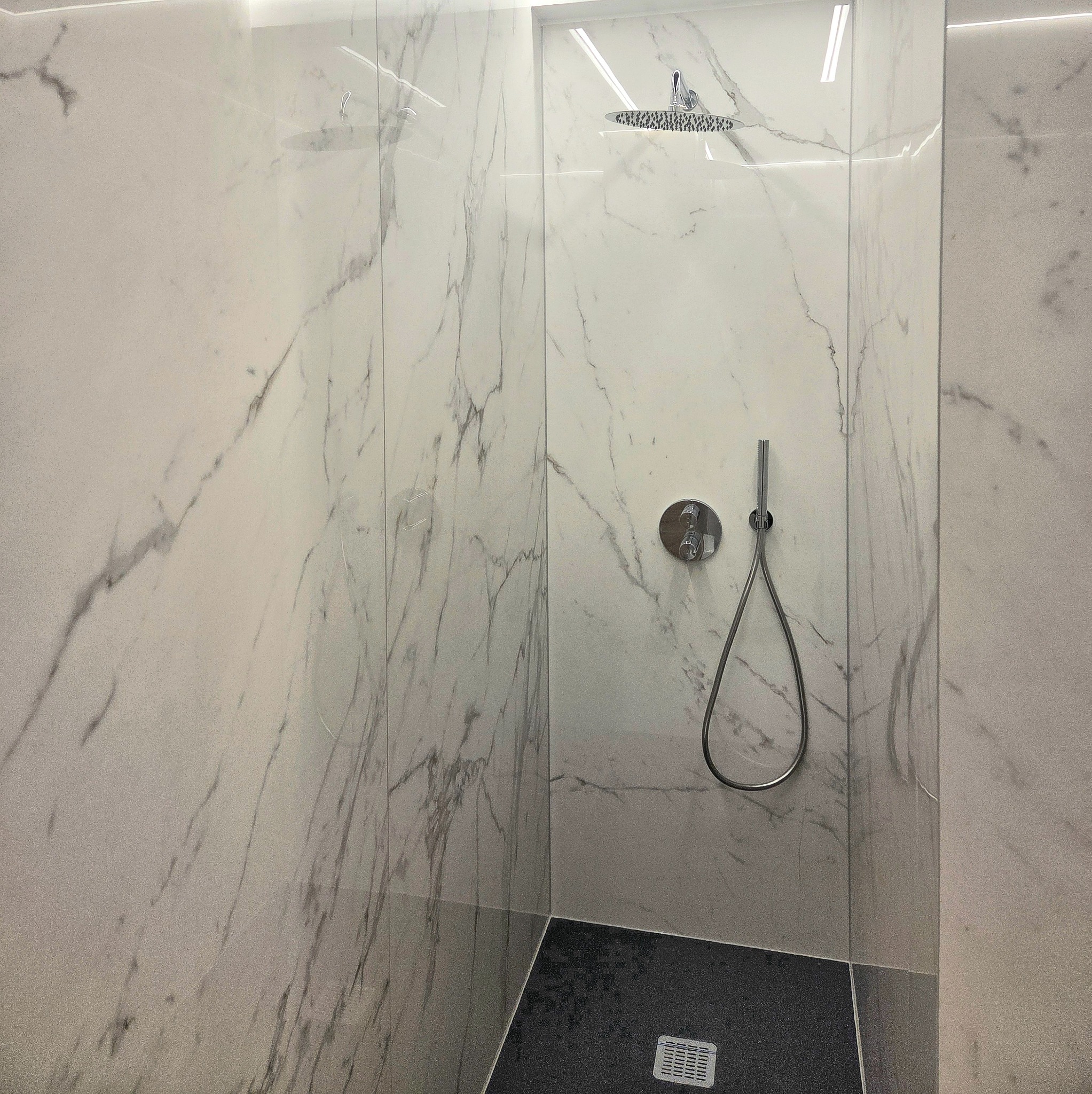 Nowoczesna kabina prysznicowa z marmurowymi płytkami, deszczownicą i szklaną ścianką. Uwagę przykuwa kontrast czarnej podłogi i jasnych ścian.