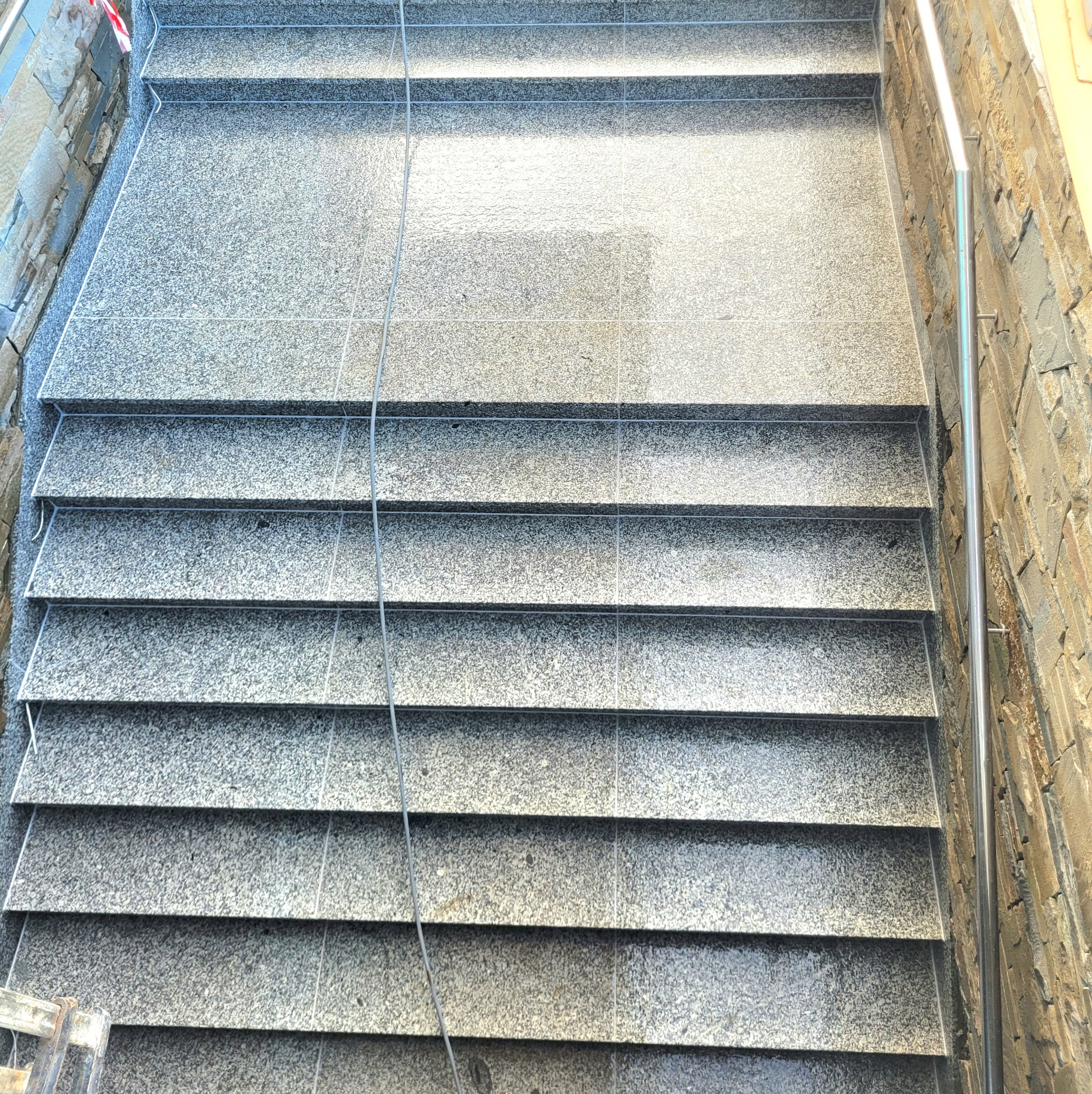 Granitowe schody zewnętrzne z metalową barierką, wykończone kamieniem elewacyjnym. Widok z góry, podkreślający fakturę i precyzję wykonania.