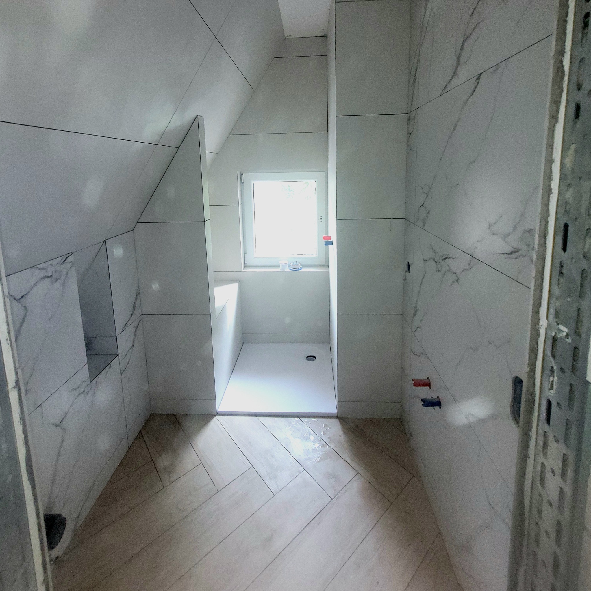 Jasna łazienka na poddaszu z kabiną prysznicową i oknem. Ściany i podłoga wyłożone płytkami imitującymi marmur i drewno. Widoczne elementy instalacji.