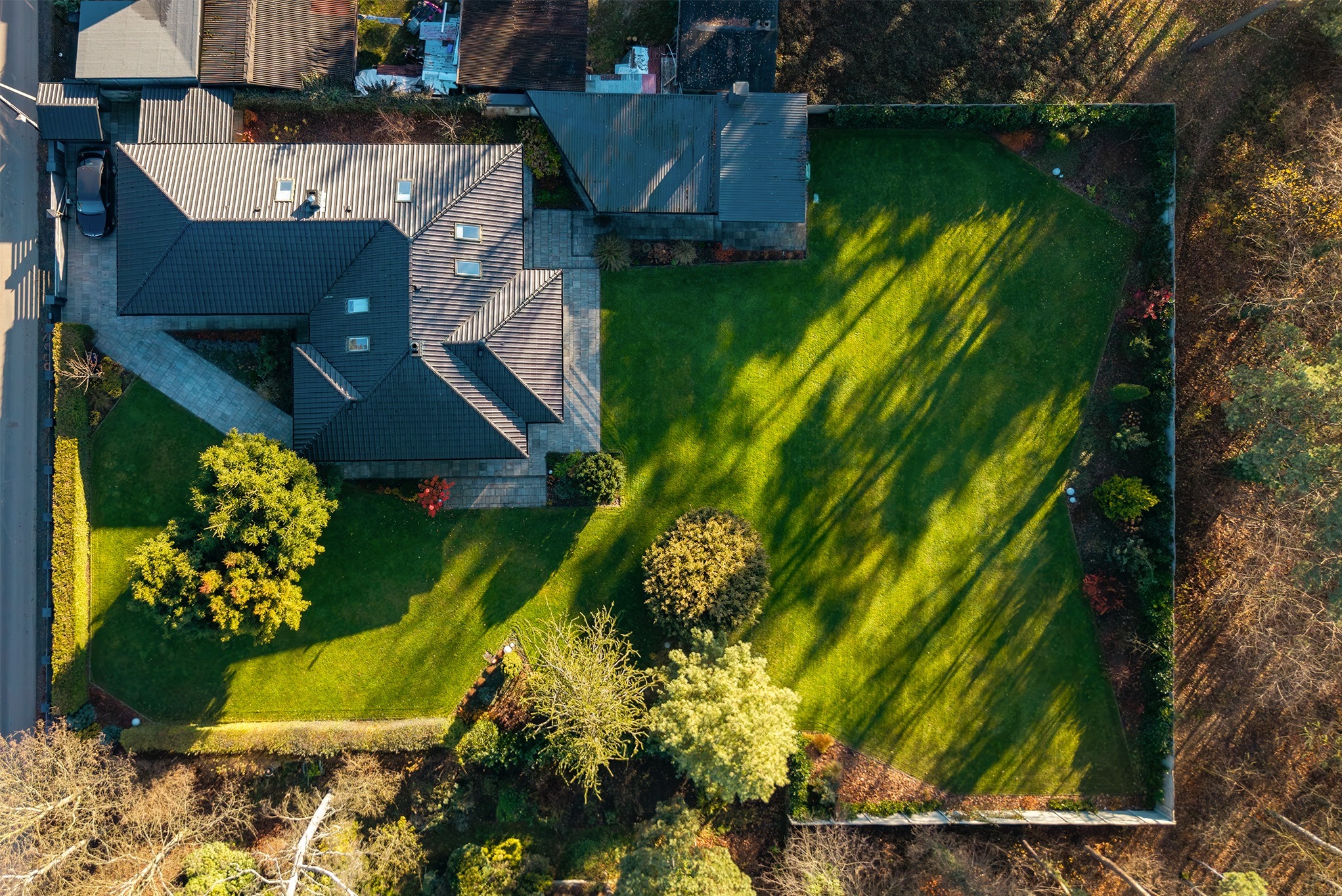 Zdjęcie z drona przedstawia dom z ciemnym dachem i zadbanym, kwadratowym trawnikiem oświetlonym promieniami słońca. Widok z góry ukazuje geometrię budynku i ogrodu.