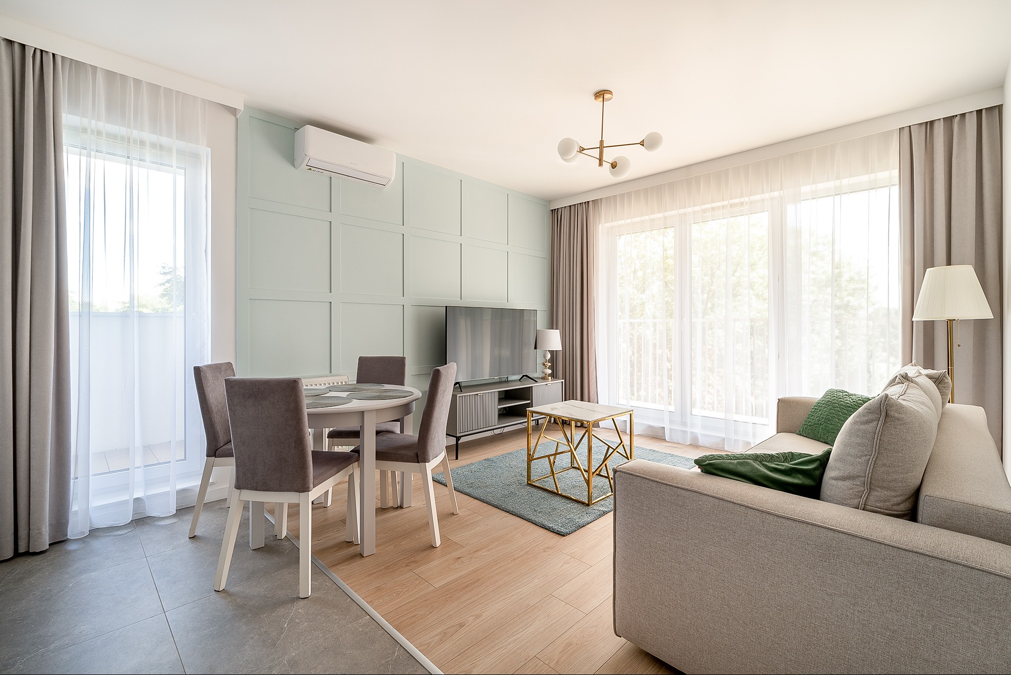 Jasne wnętrze salonu z okrągłym stołem, szarą sofą i telewizorem na tle ściany w pastelowej zieleni. Nowoczesny design, naturalne światło wpadające przez okna.