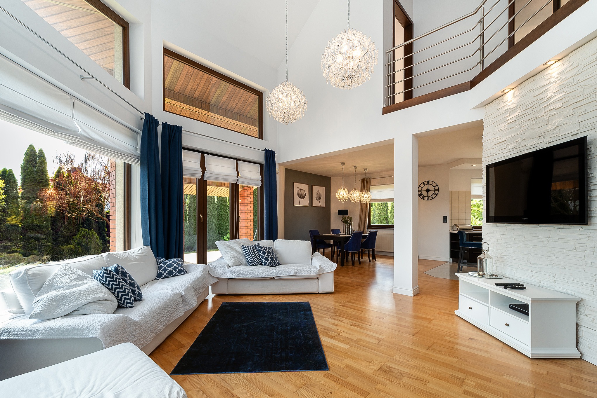 Jasne wnętrze domu z wysokim sufitem, białymi sofami z wzorzystymi poduszkami, drewnianą podłogą i widokiem na ogród przez duże okno. Dwie kryształowe lampy.