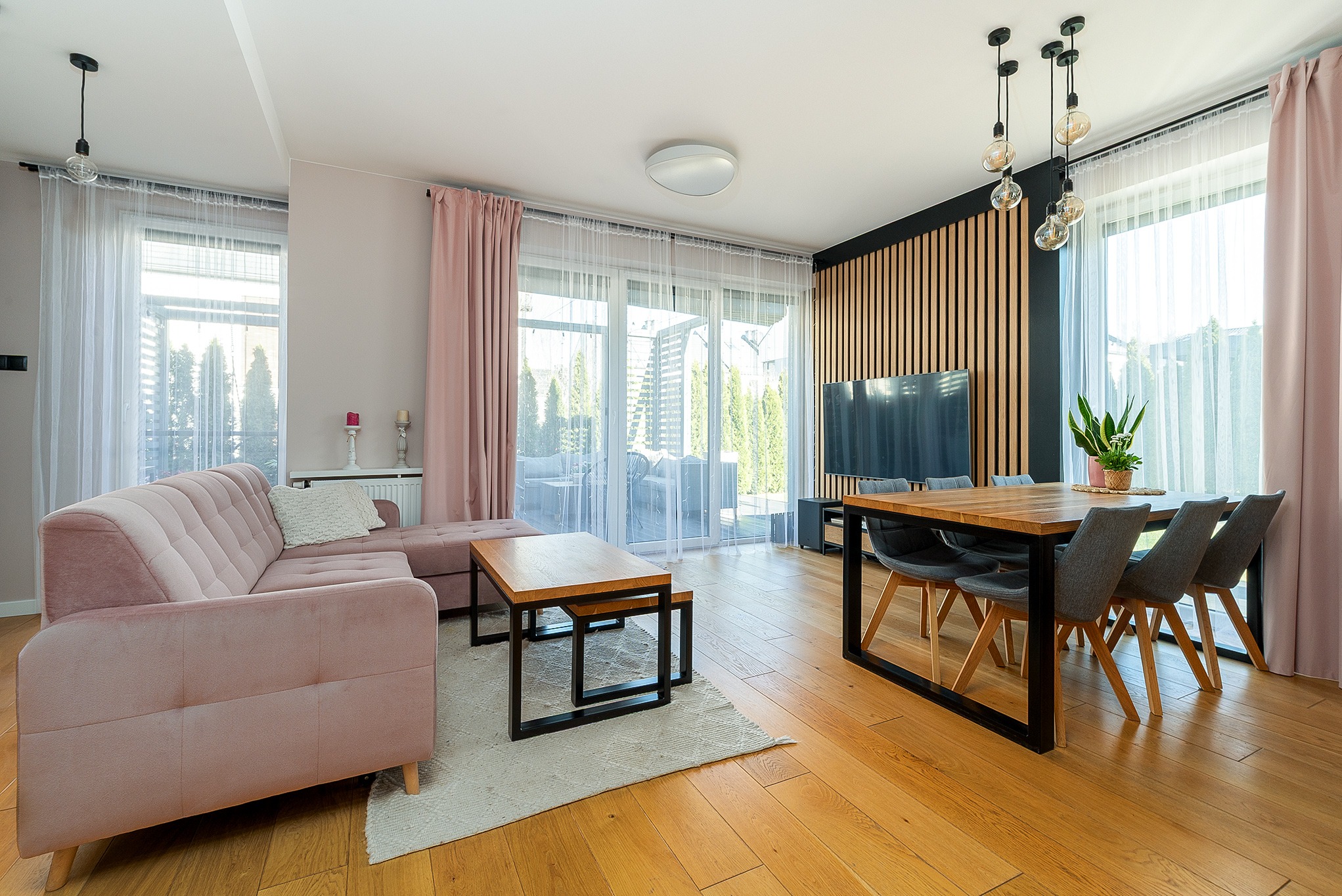 Jasne wnętrze salonu z drewnianą podłogą, różową sofą i stołem jadalnym. Ściana z drewnianymi panelami i nowoczesne lampy wiszące. Duże okna z widokiem na ogród.