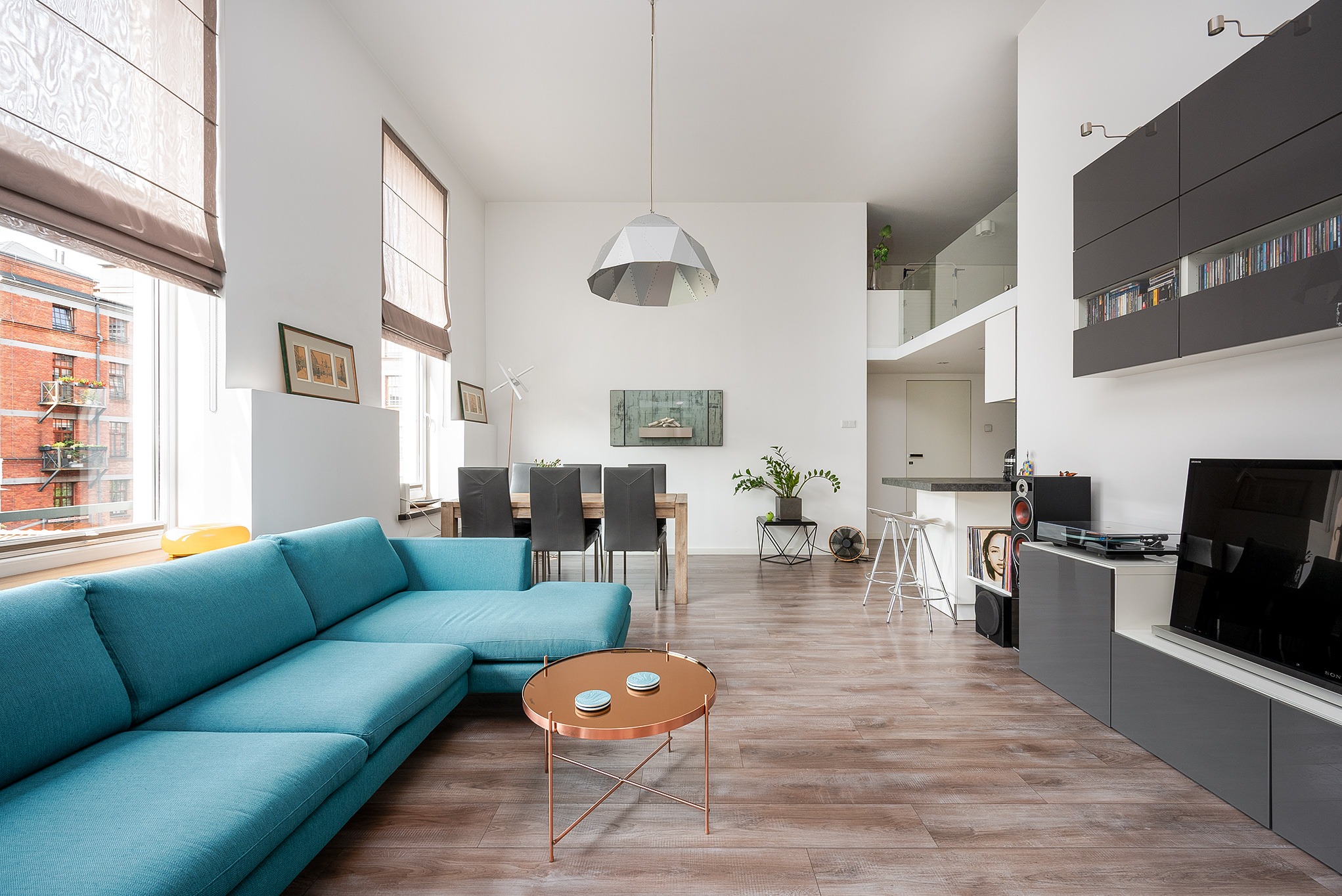 Przestronny salon w Łodzi z turkusową sofą narożną, okrągłym stolikiem kawowym i drewnianą podłogą. Widok na dwupoziomowe wnętrze z elementami nowoczesnego designu.