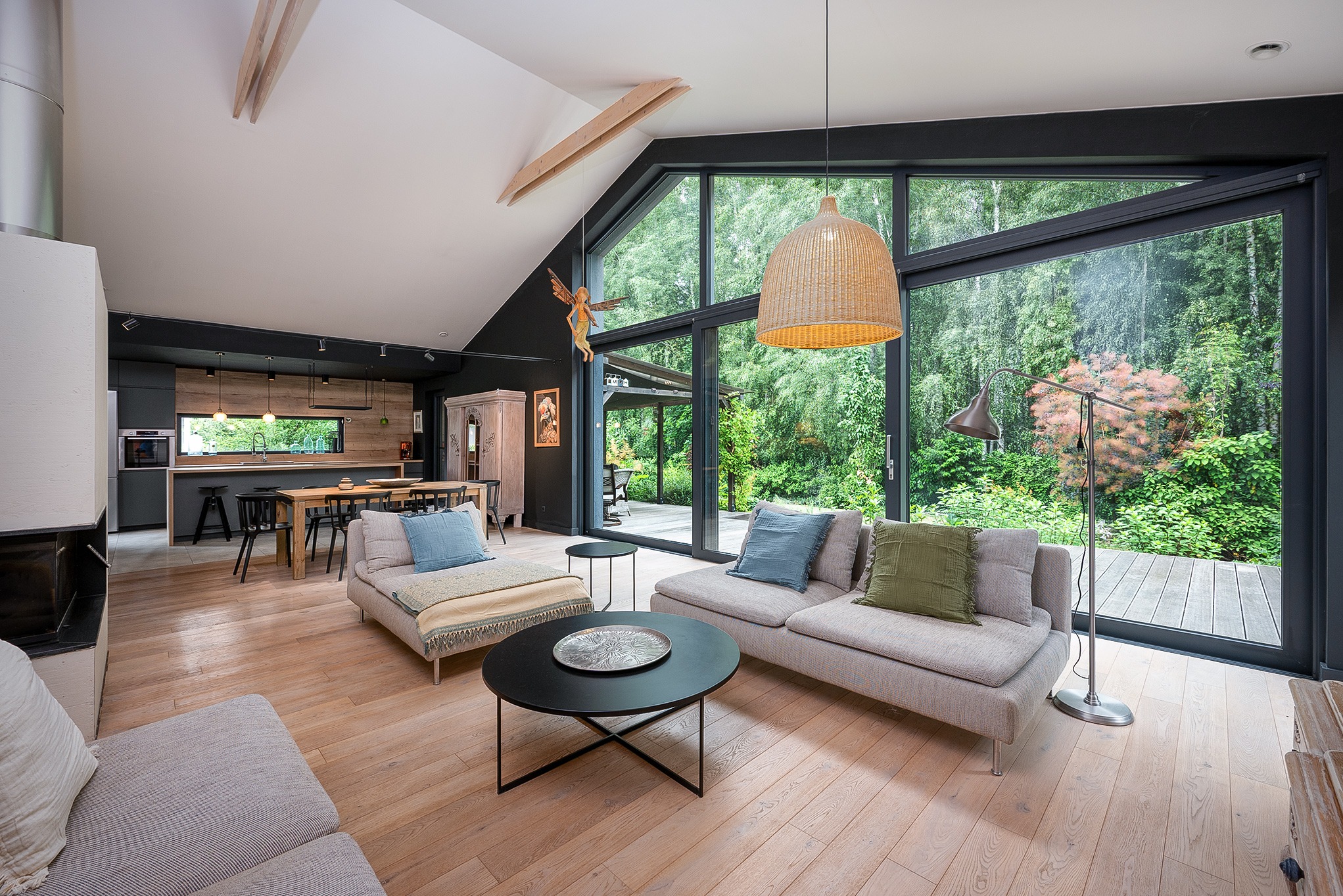 Jasne, przestronne wnętrze domu z dużym oknem na las. Dwie sofy, stolik kawowy i widok na kuchnię. W tle zieleń drzew i krzewów. Wnętrze urządzone w nowoczesnym stylu.