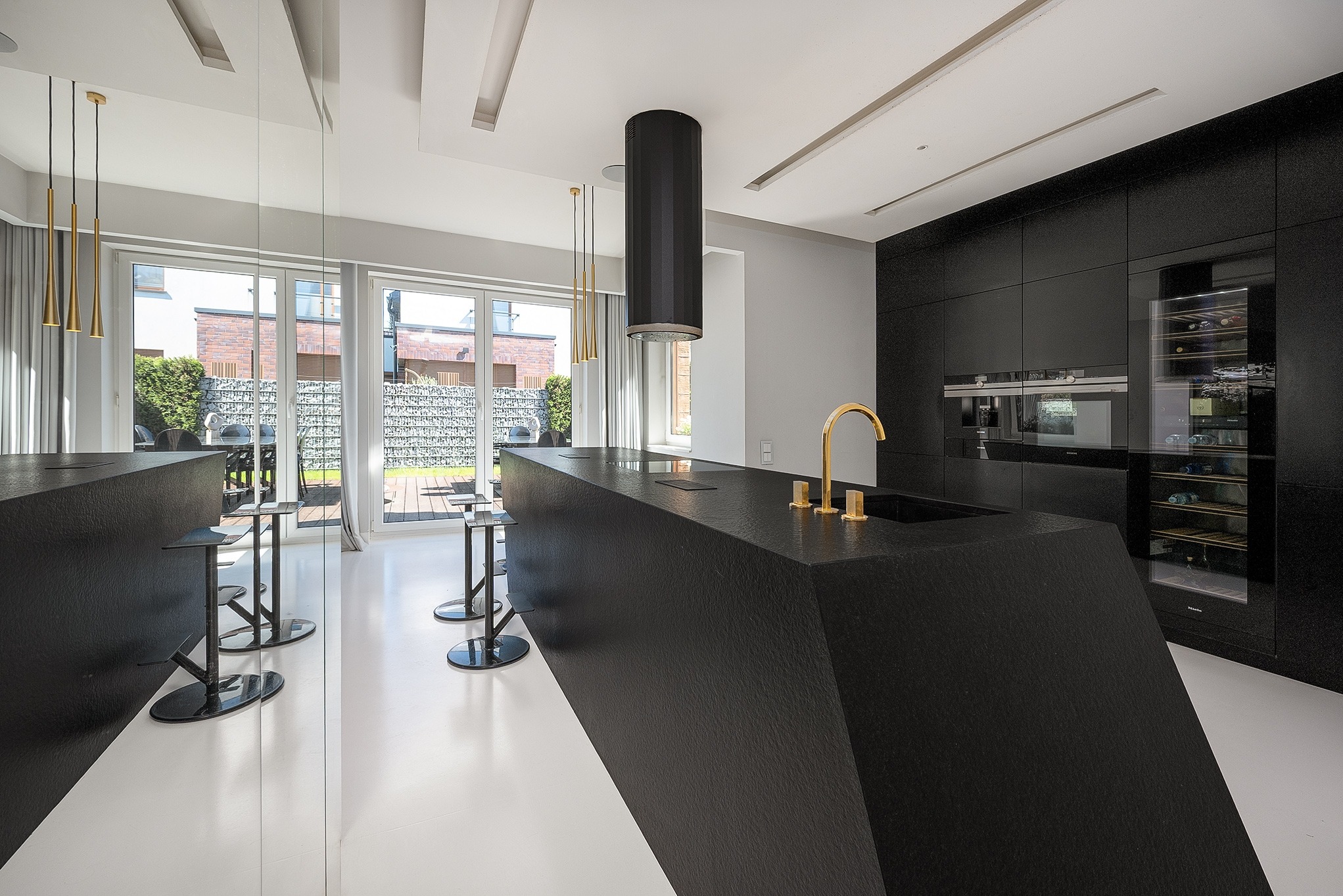 Nowoczesna kuchnia z czarną wyspą i złotą armaturą. Minimalistyczny design, biała podłoga, duże okna z widokiem na ogród. Eleganckie oświetlenie.