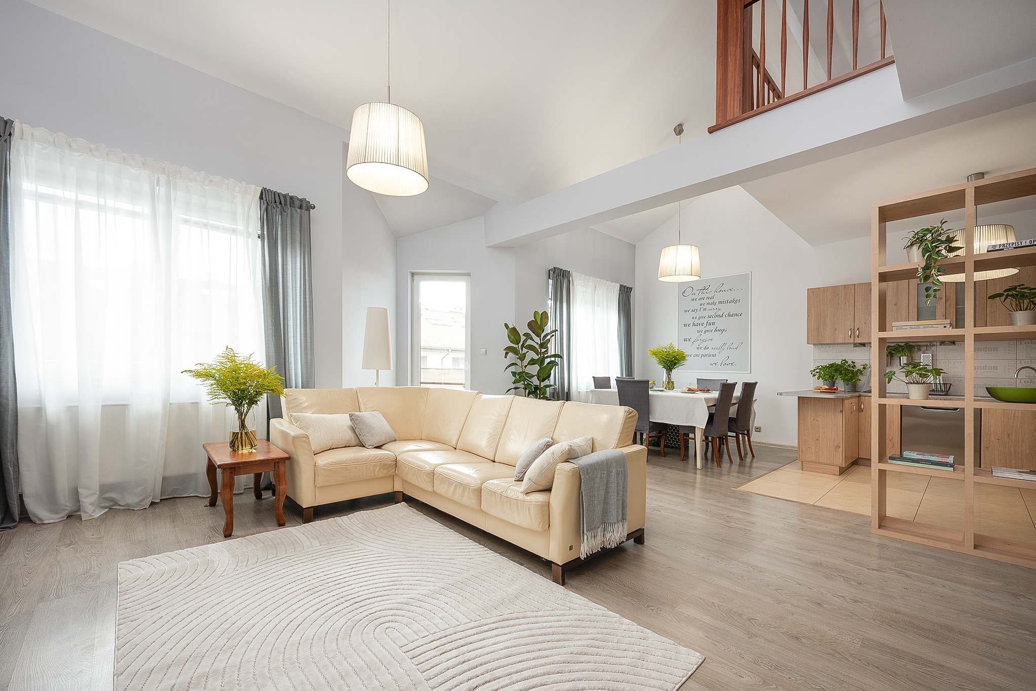 Jasne wnętrze domu z beżową sofą narożną, wzorzystym dywanem i drewnianymi elementami. Widok na jadalnię i schody na antresolę. Neutralna kolorystyka i naturalne światło.