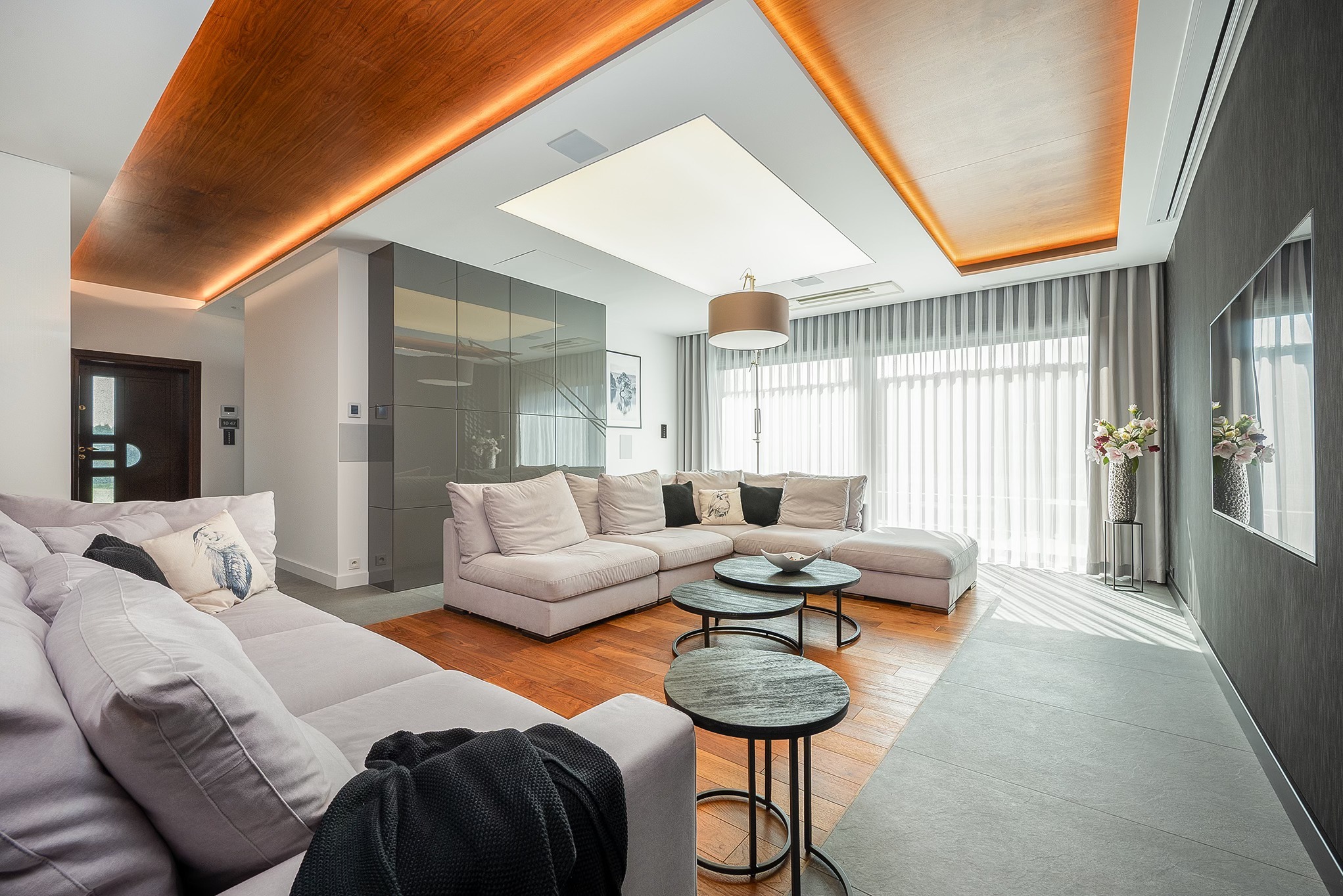 Jasny salon z drewnianą podłogą i sufitem, szarą sofą narożną, okrągłymi stolikami kawowymi i dużym oknem z zasłonami. Nowoczesne oświetlenie sufitowe.