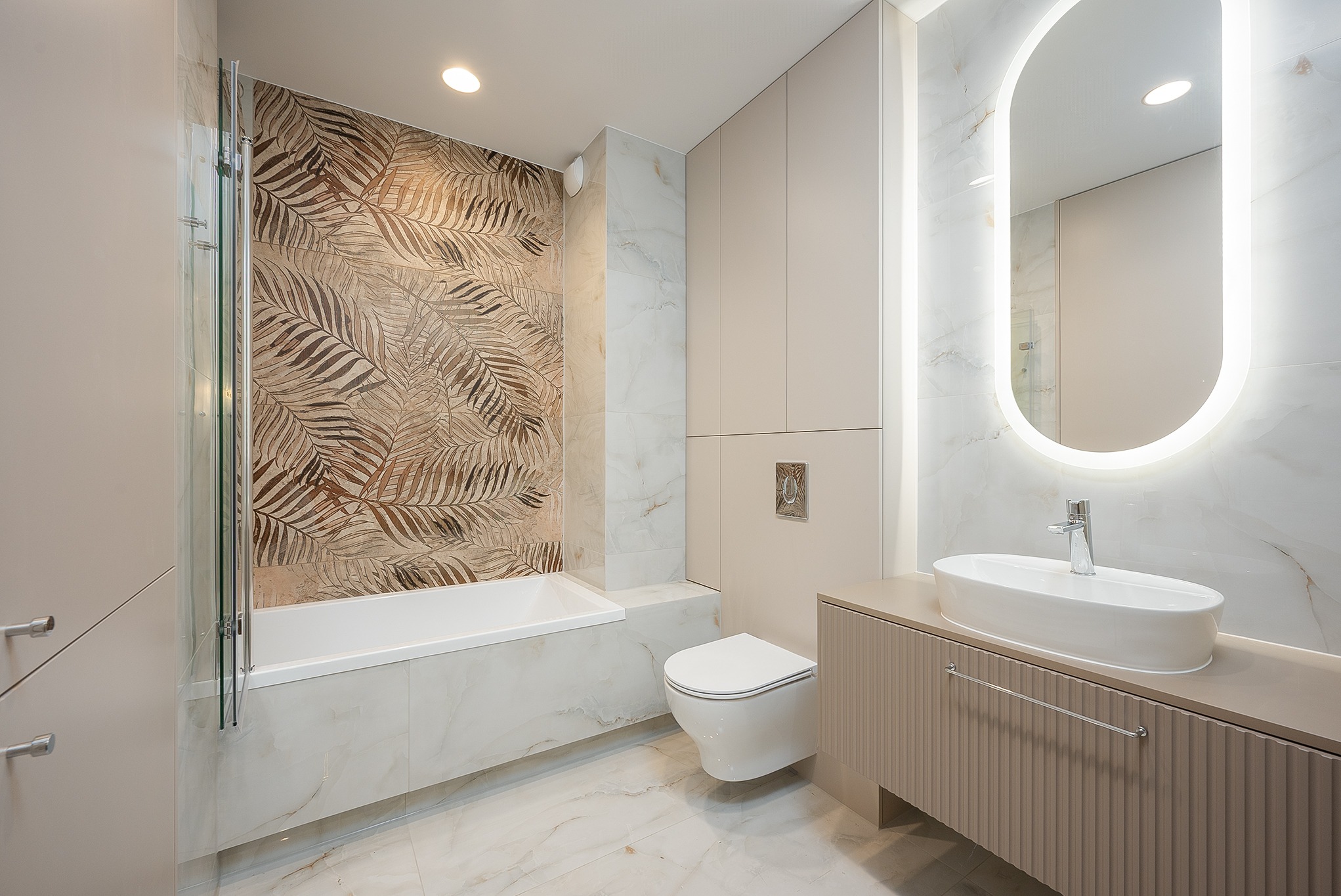 Nowoczesna łazienka w beżach: wanna z parawanem, toaleta podtynkowa, umywalka nablatowa z szafką i podświetlane lustro owalne. Akcent: ściana z motywem liści.