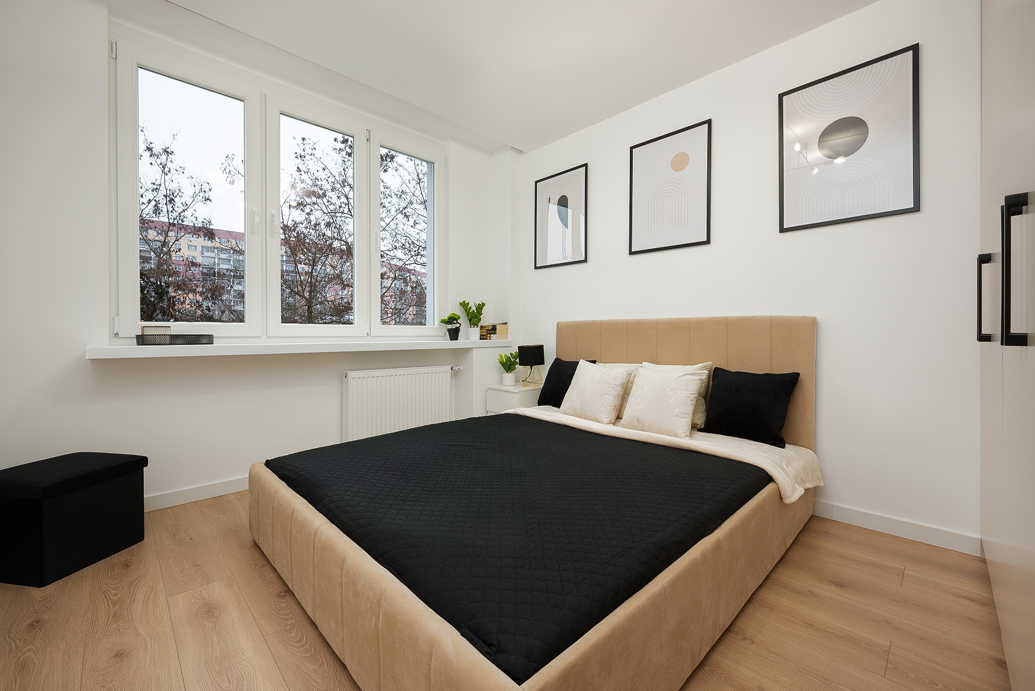 Jasna sypialnia z beżowym łóżkiem, czarną narzutą i poduszkami. Na ścianie grafiki w czarnych ramkach. Widok z okna na budynek. Minimalistyczny wystrój wnętrza.