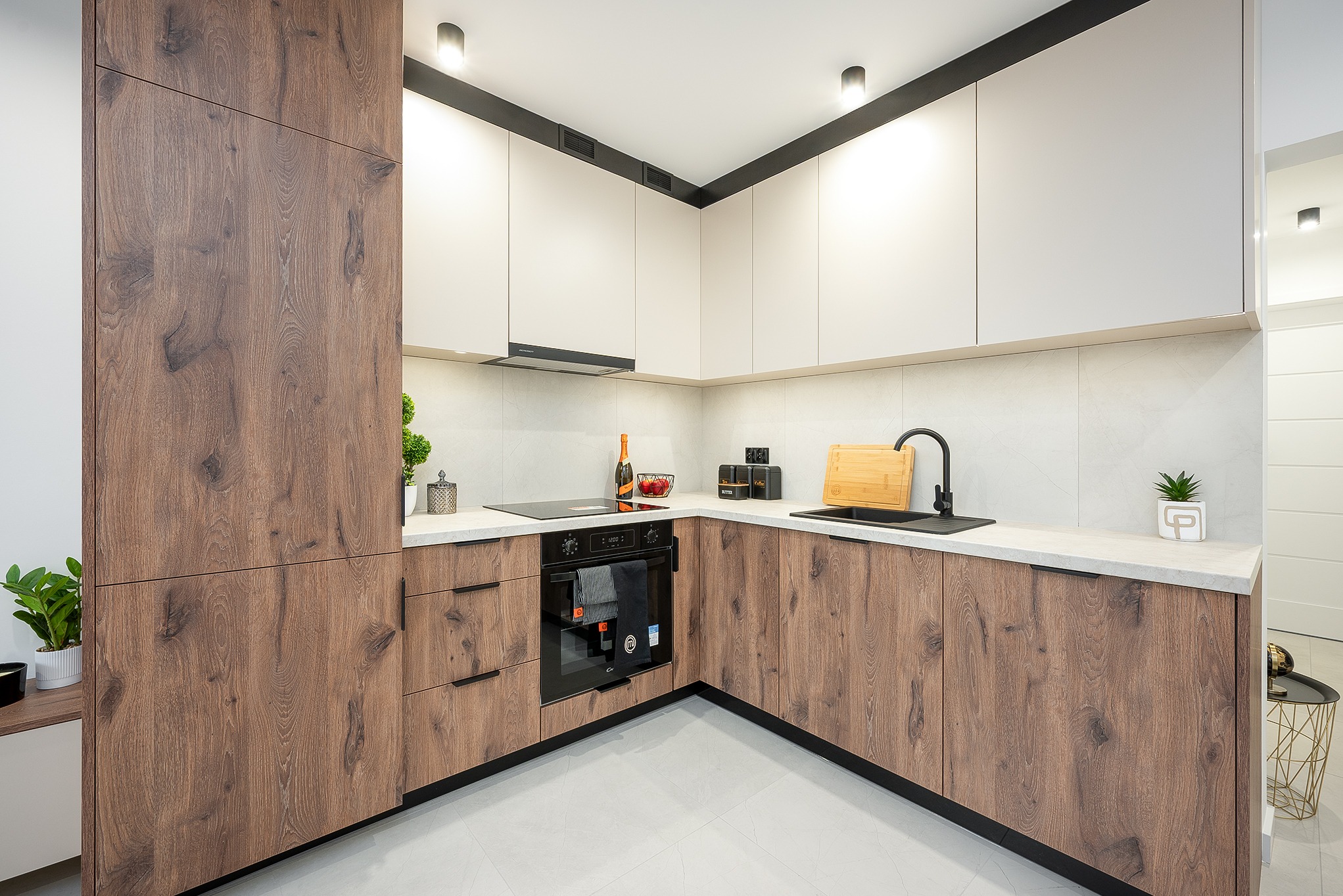 Nowoczesna kuchnia w kształcie litery L z drewnianymi frontami szafek i białymi górnymi szafkami, czarnymi akcentami i roślinami doniczkowymi.