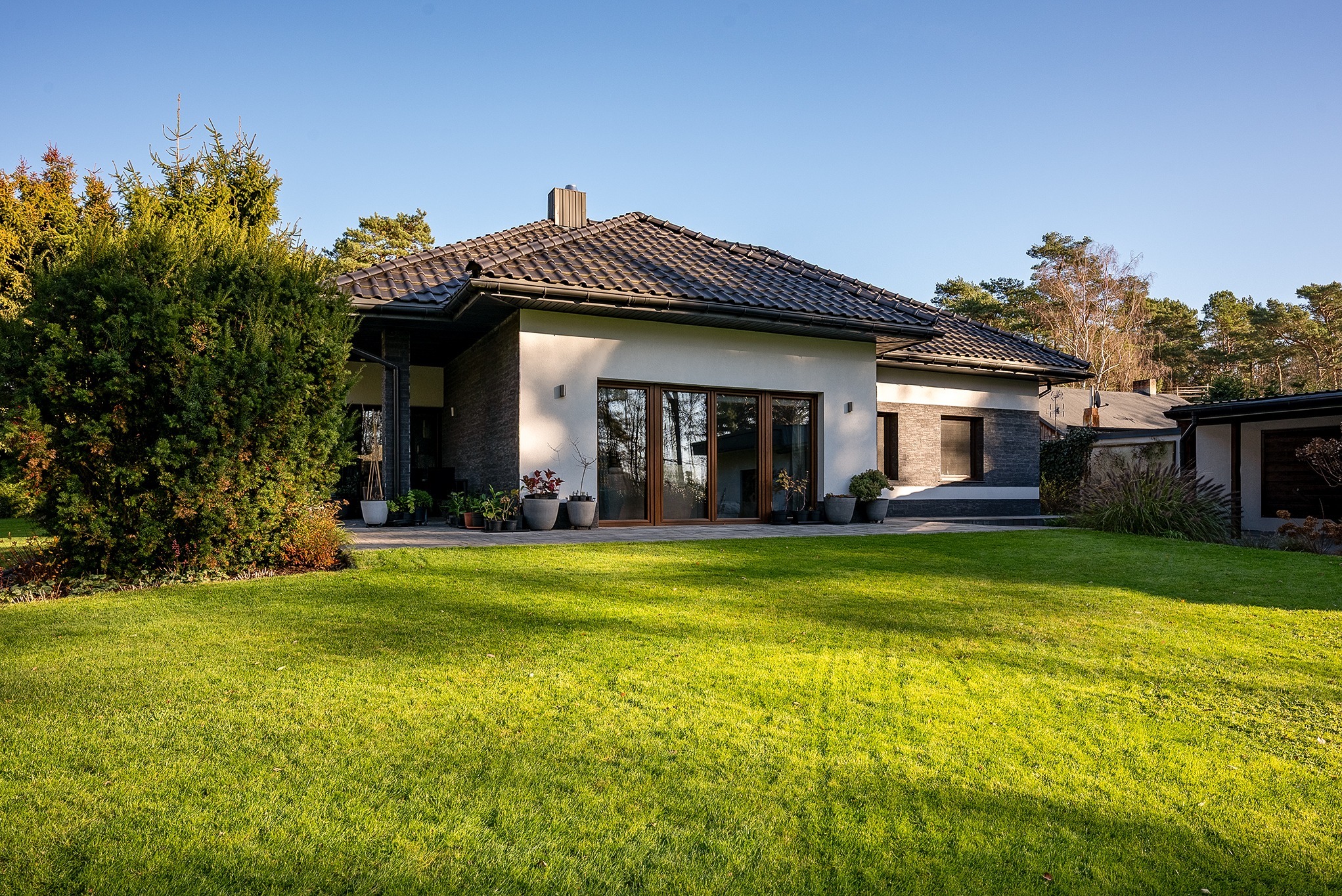 Elewacja domu jednorodzinnego z dużym trawnikiem, widok z zewnątrz. Nowoczesna architektura z elementami kamienia i drewna. Uwagę przykuwają duże okna i zadbany ogród.