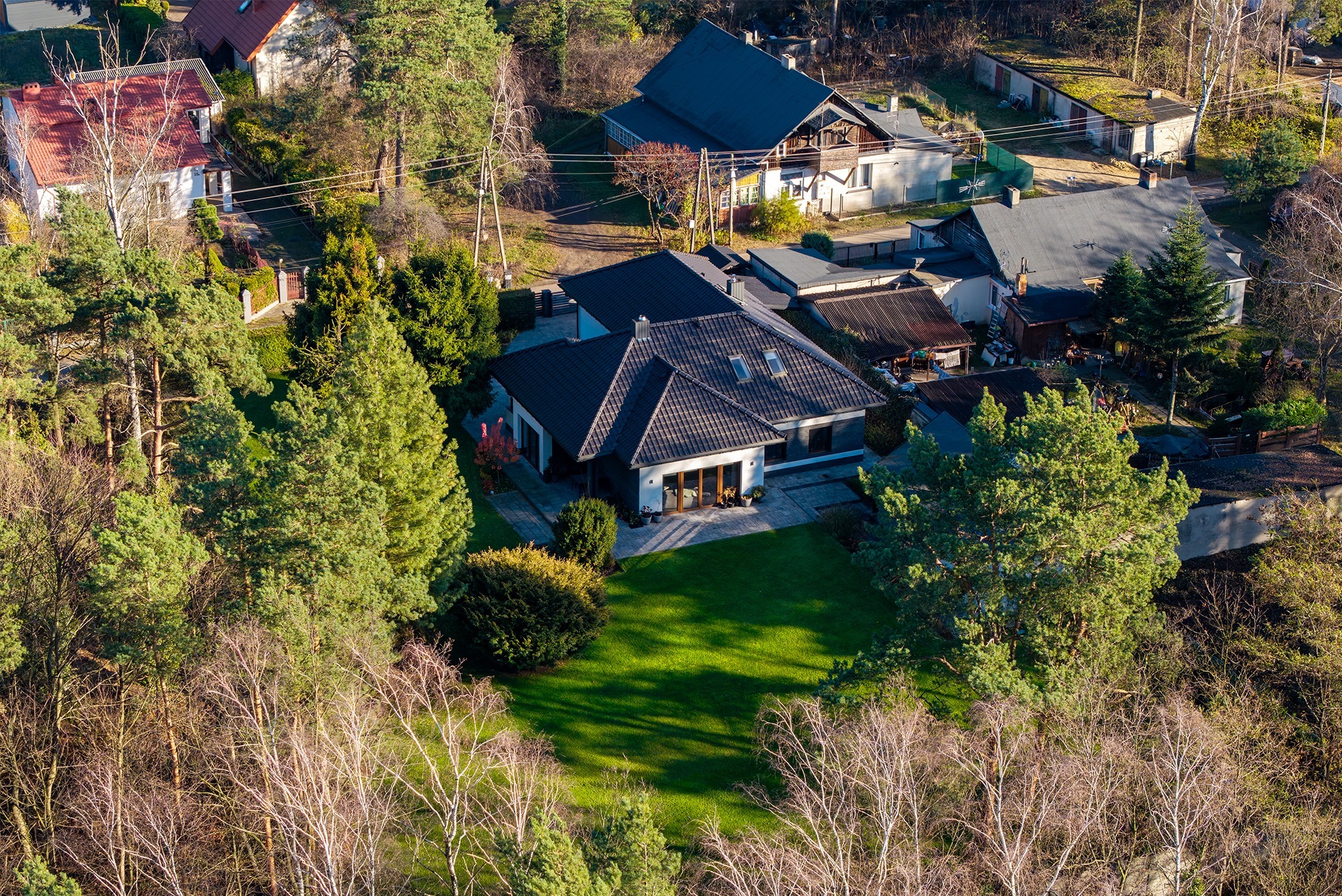 Zdjęcie z lotu ptaka przedstawia nowoczesny dom z ciemnym dachem i dużym ogrodem, otoczony drzewami. Widoczne są sąsiednie budynki i elementy krajobrazu.
