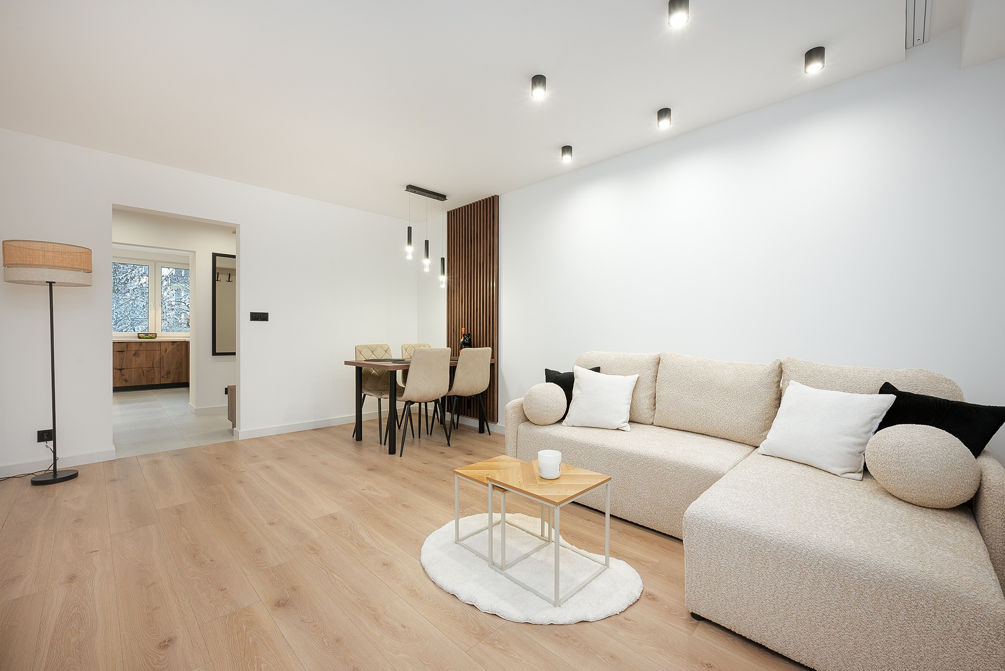 Jasne wnętrze: beżowa sofa z poduszkami, stolik na dywaniku, stół z krzesłami i lampa podłogowa. Minimalistyczny design z drewnianą podłogą i białymi ścianami.