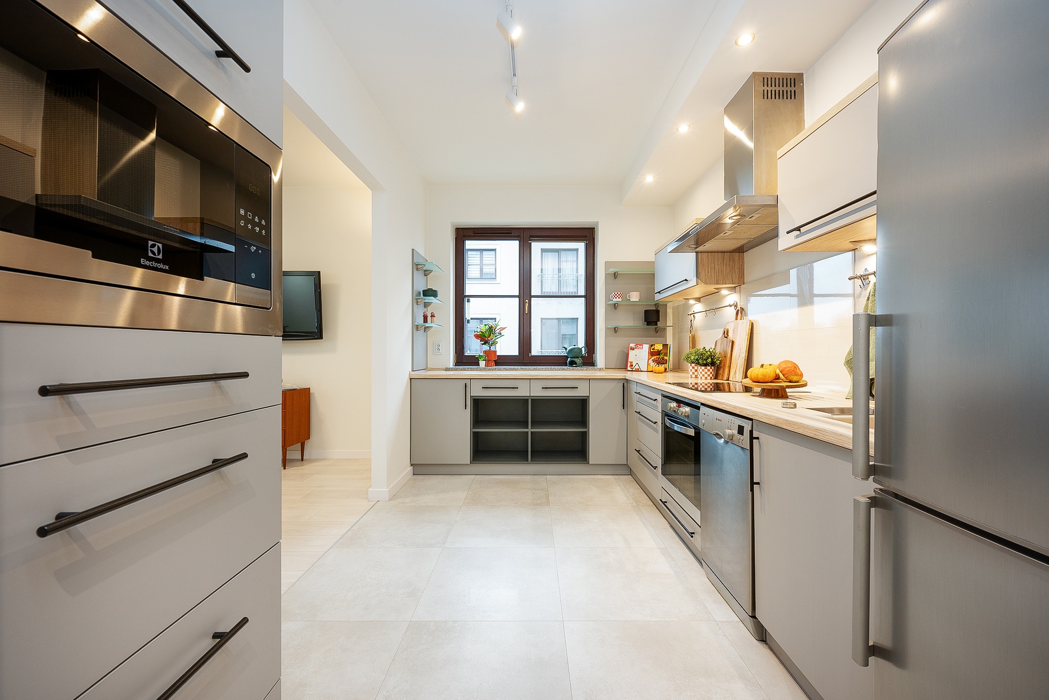 Jasna kuchnia w szarościach z drewnianymi akcentami. Widok na okno z roślinami i dekoracjami. Nowoczesne oświetlenie punktowe. Minimalistyczny design.