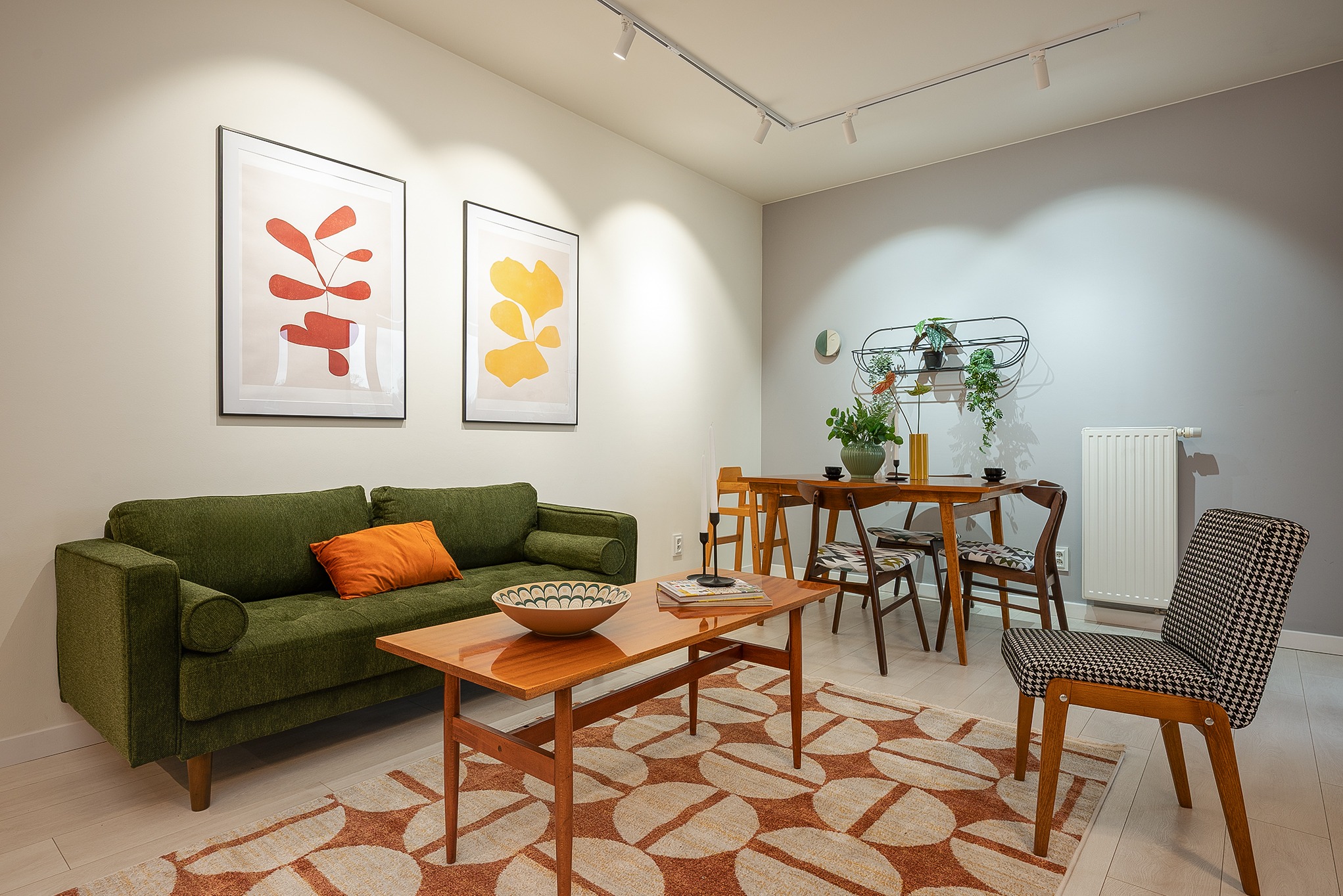 Jasne wnętrze w stylu retro: zielona sofa z pomarańczową poduszką, drewniany stół kawowy z misą, dywan w geometryczne wzory, plakaty na ścianie i oświetlenie punktowe.