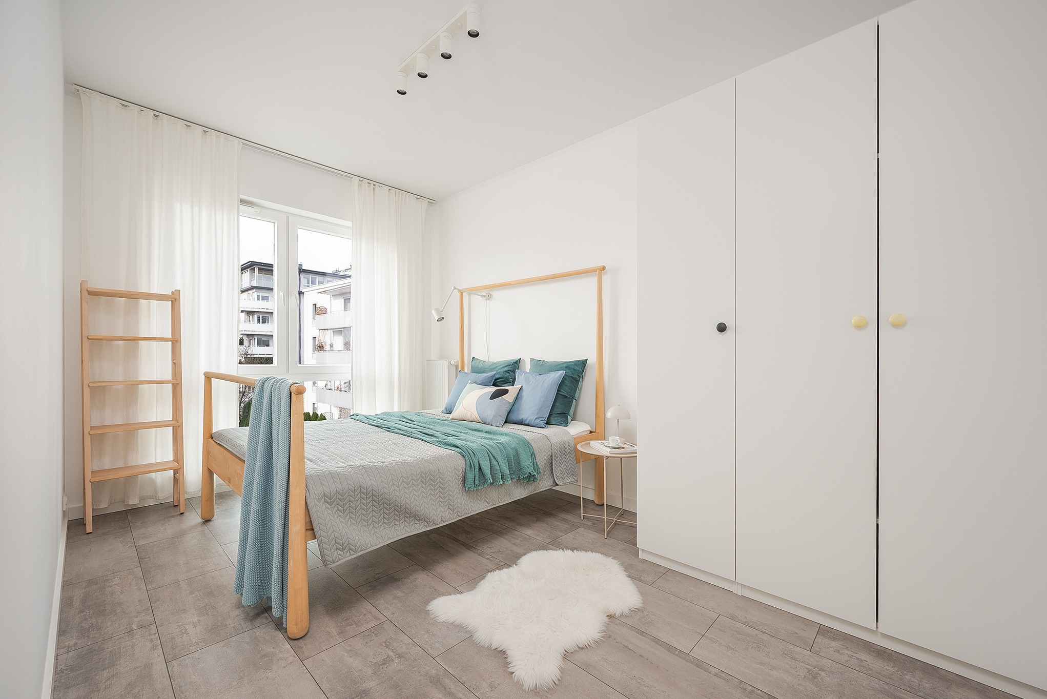 Jasna sypialnia z drewnianym łóżkiem, turkusowymi akcentami i białymi szafami w zabudowie. Minimalistyczny design, naturalne światło, dywanik z owczej skóry.