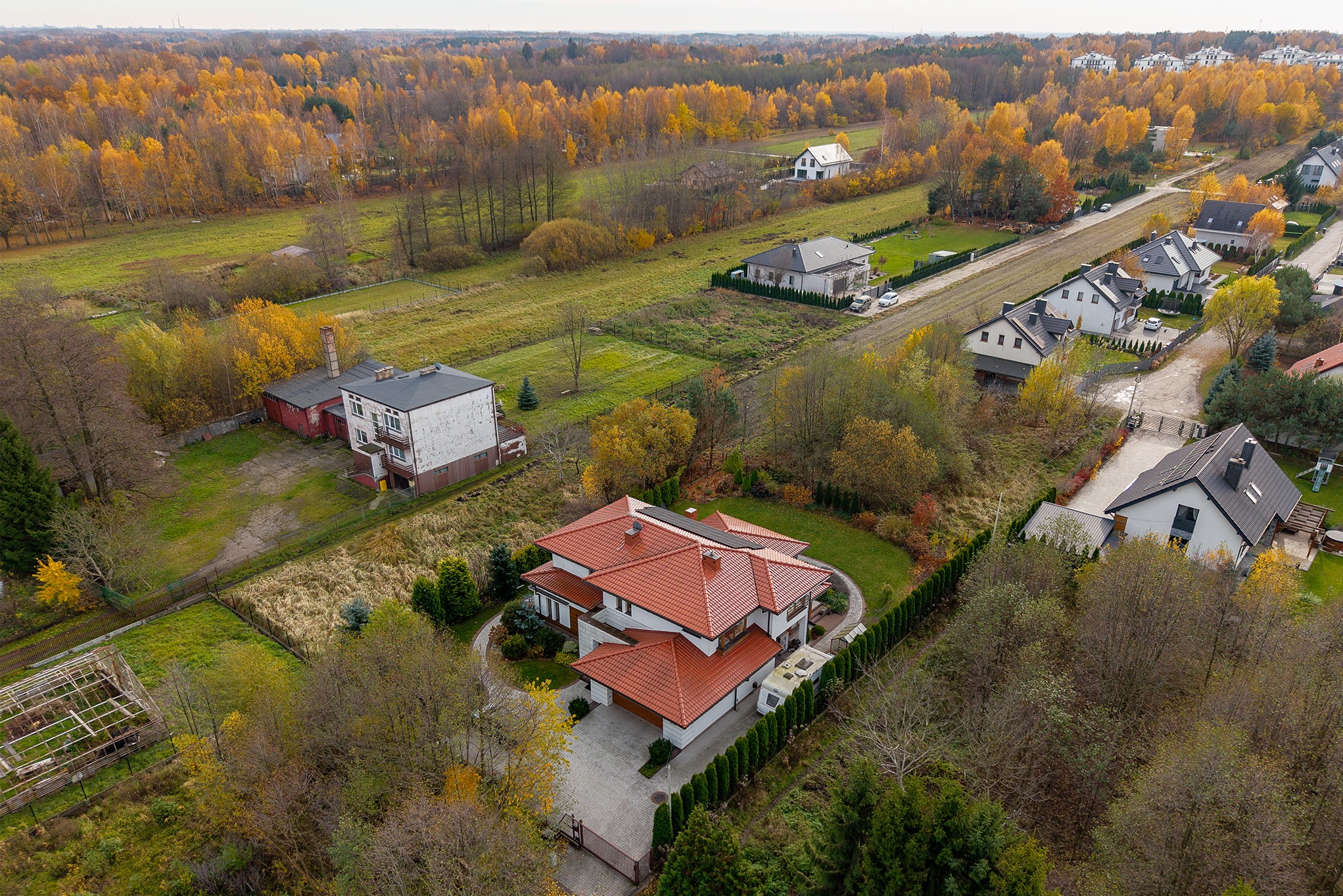 Jesienny widok z drona na nowoczesny dom z czerwoną dachówką, otoczony zielenią i drzewami w barwach jesieni. Ujęcie z góry ukazuje architekturę i krajobraz.
