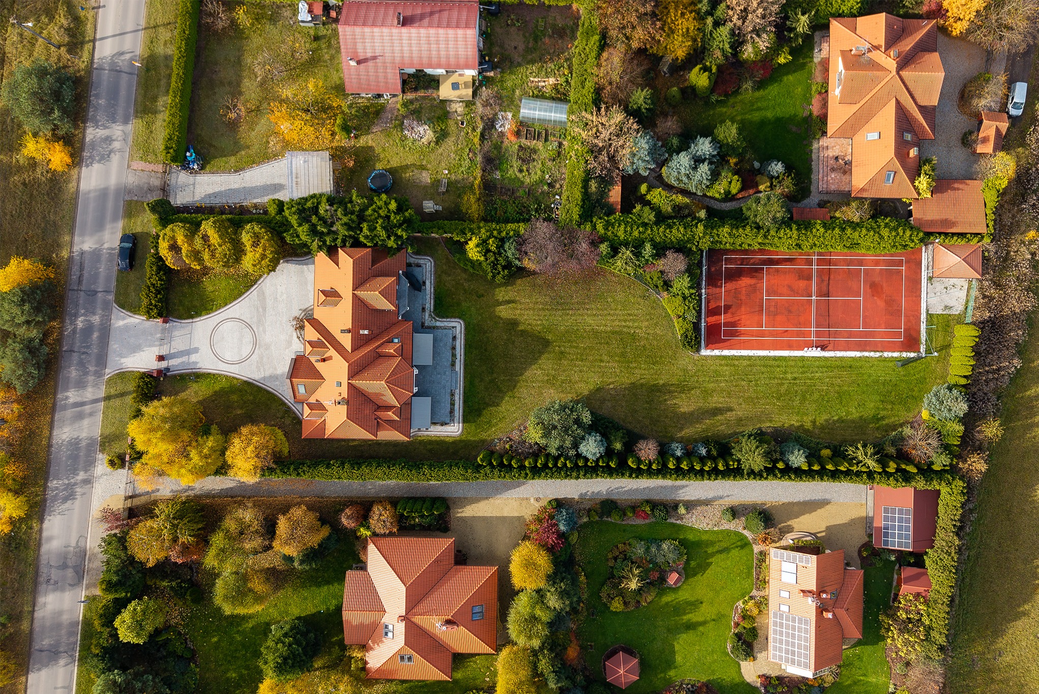Widok z drona na rezydencje z kortem tenisowym, otoczone zielenią i jesiennymi drzewami. Czerwone dachy domów kontrastują z zielenią trawników i barwnymi drzewami.