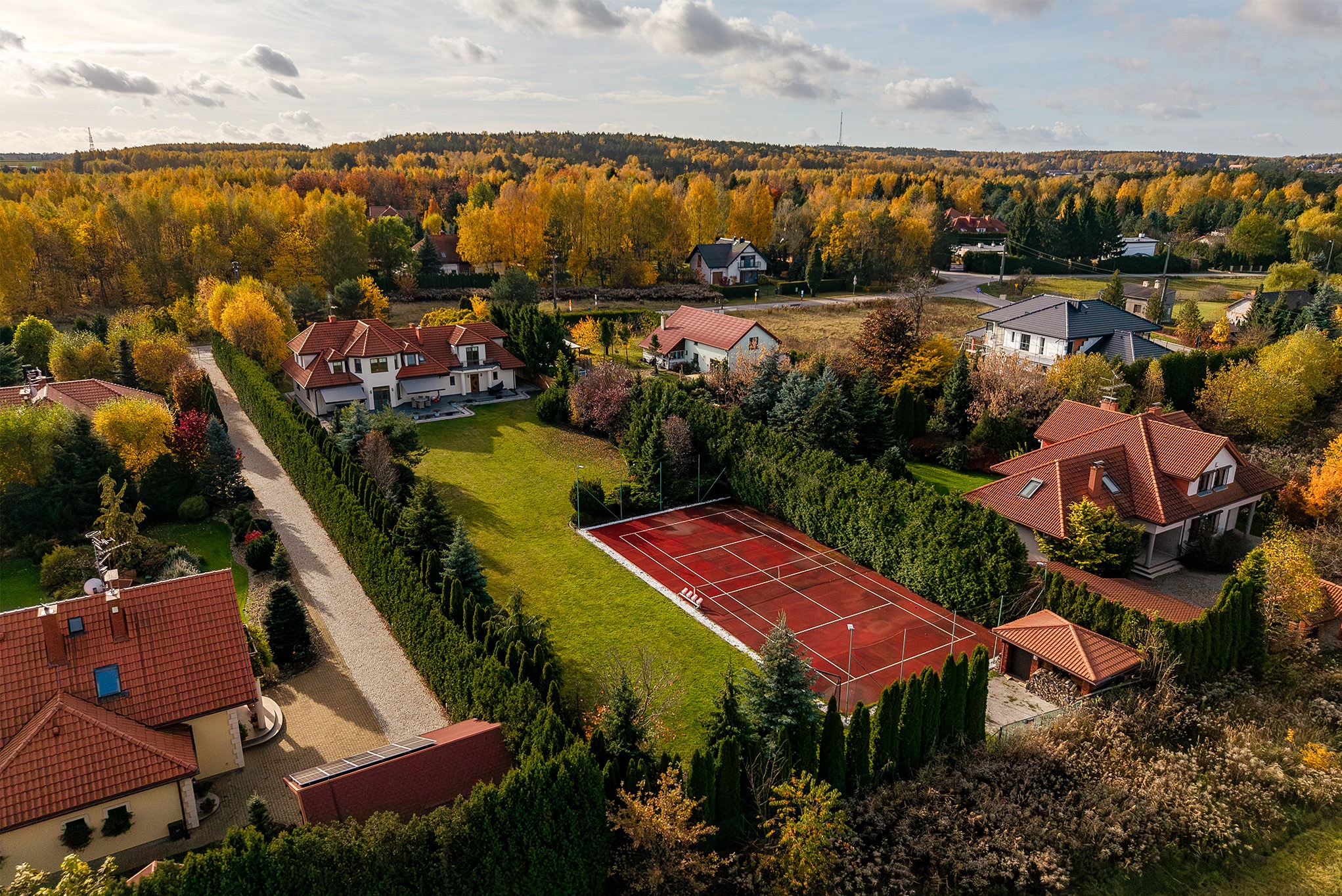 Jesienny widok z lotu ptaka na domy jednorodzinne z czerwonymi dachami i kort tenisowy otoczony zielenią. Drzewa w odcieniach żółci i pomarańczy. Słoneczny dzień.