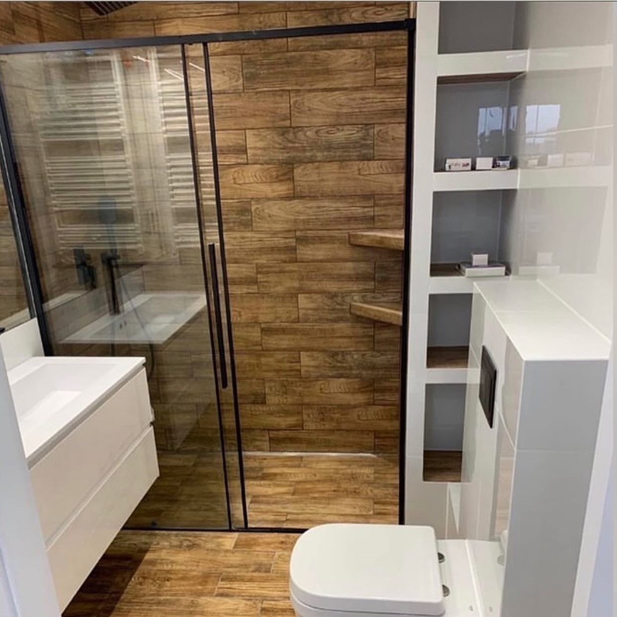 Nowoczesna łazienka z kabiną prysznicową typu walk-in, wykończona płytkami imitującymi drewno i białymi elementami. Widoczne umywalka, toaleta i półki w ścianie.