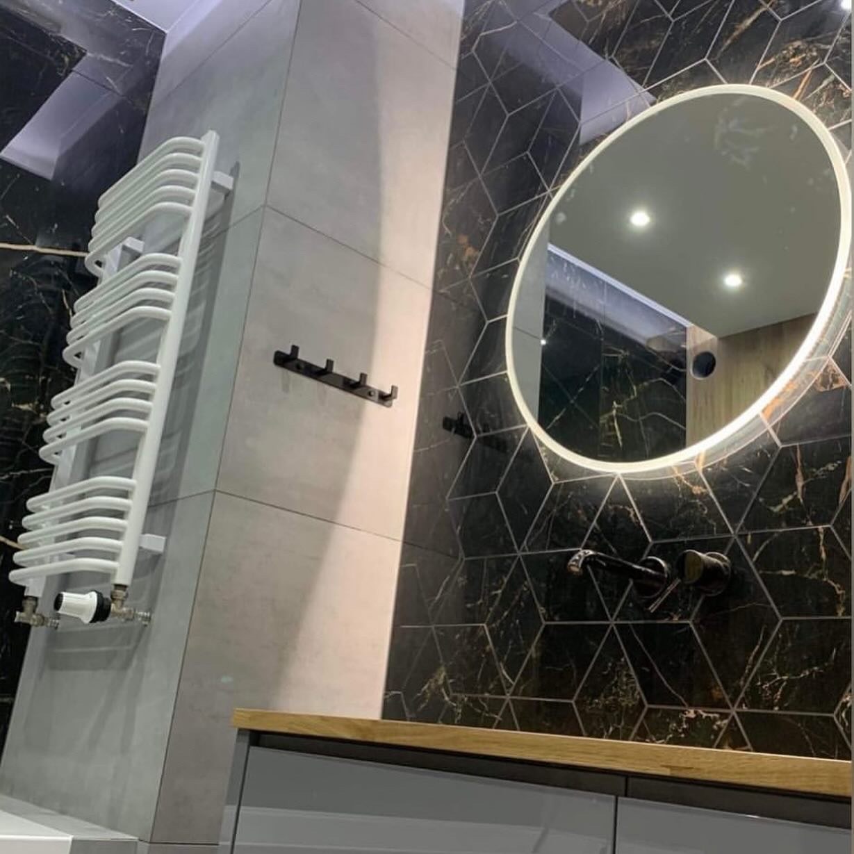 Nowoczesna łazienka z czarną mozaiką, podświetlanym lustrem i białym grzejnikiem drabinkowym. Minimalistyczne akcesoria i drewniany blat dodają elegancji.