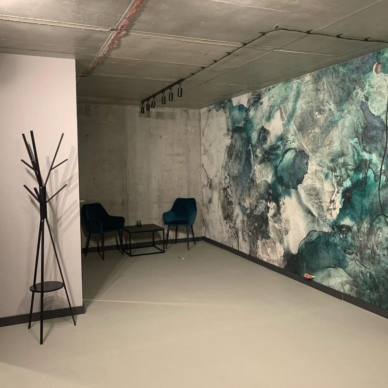 Wykończone wnętrze pokoju z abstrakcyjną tapetą w odcieniach zieleni i szarości, betonowym sufitem z oświetleniem szynowym i minimalistycznym wieszakiem na ubrania.