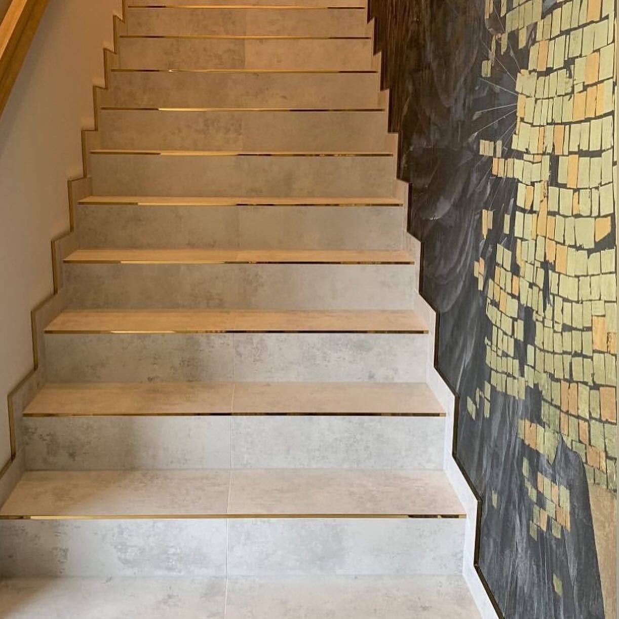 Wykończone schody z płytkami w odcieniach szarości, z delikatnymi złotymi listwami. Na ścianie widoczna dekoracyjna tapeta w odcieniach złota i szarości.