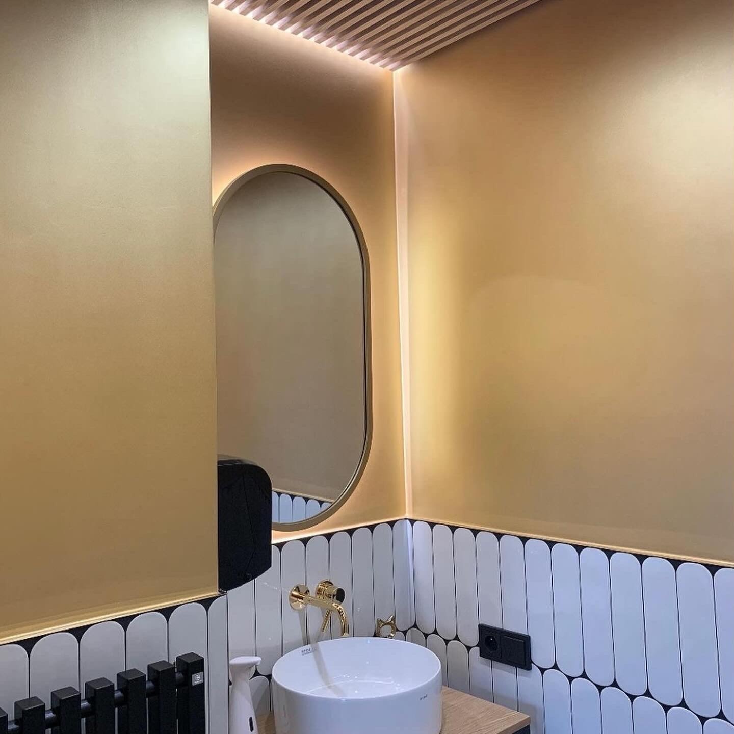 Złota łazienka z owalnym lustrem, białymi płytkami w czarne obramowania i czarnym grzejnikiem. Nowoczesny design z oświetleniem LED na suficie.