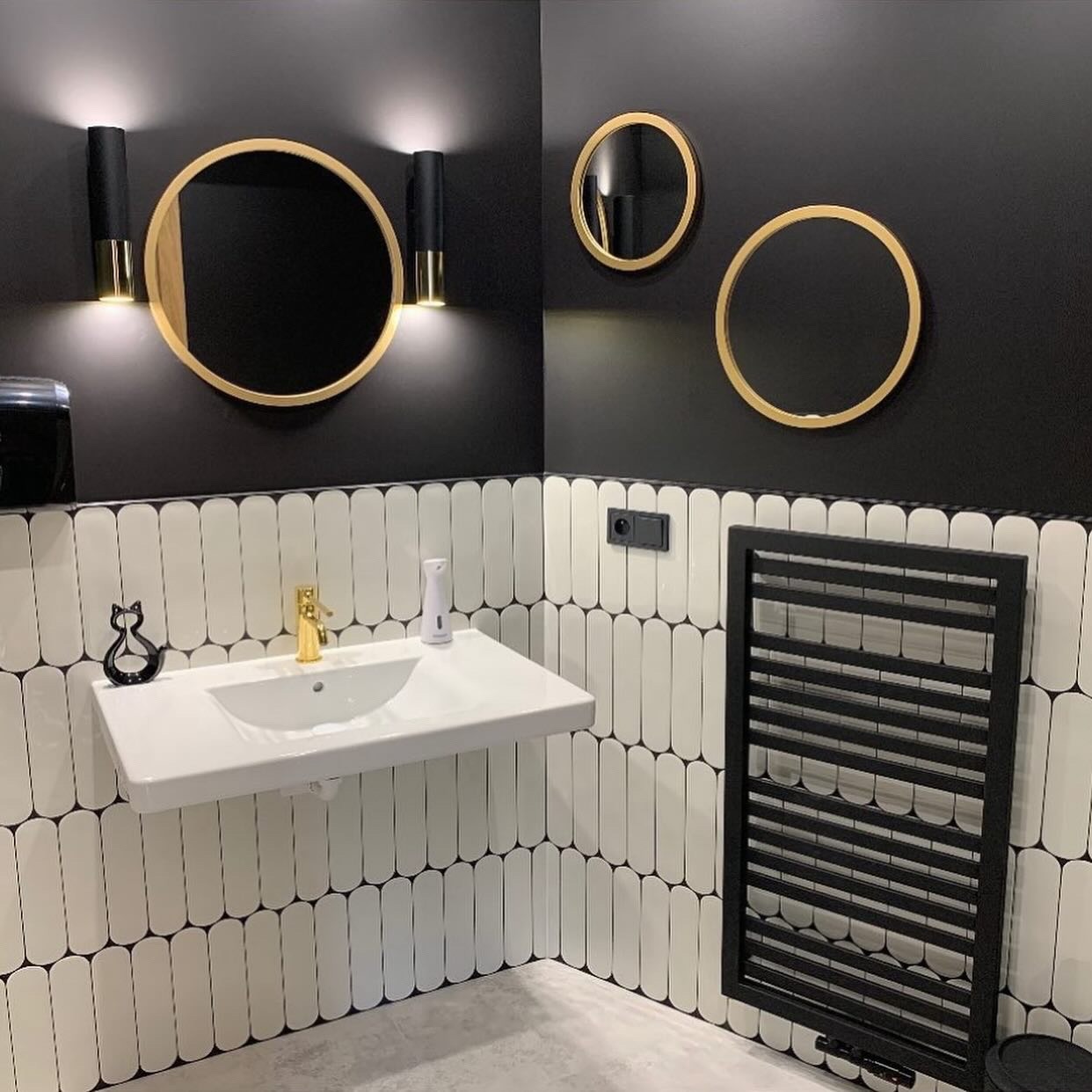 Nowoczesna łazienka w czerni i bieli z okrągłymi lustrami w złotych ramach, podświetlanymi kinkietami i umywalką na tle płytek w kształcie migdałów.