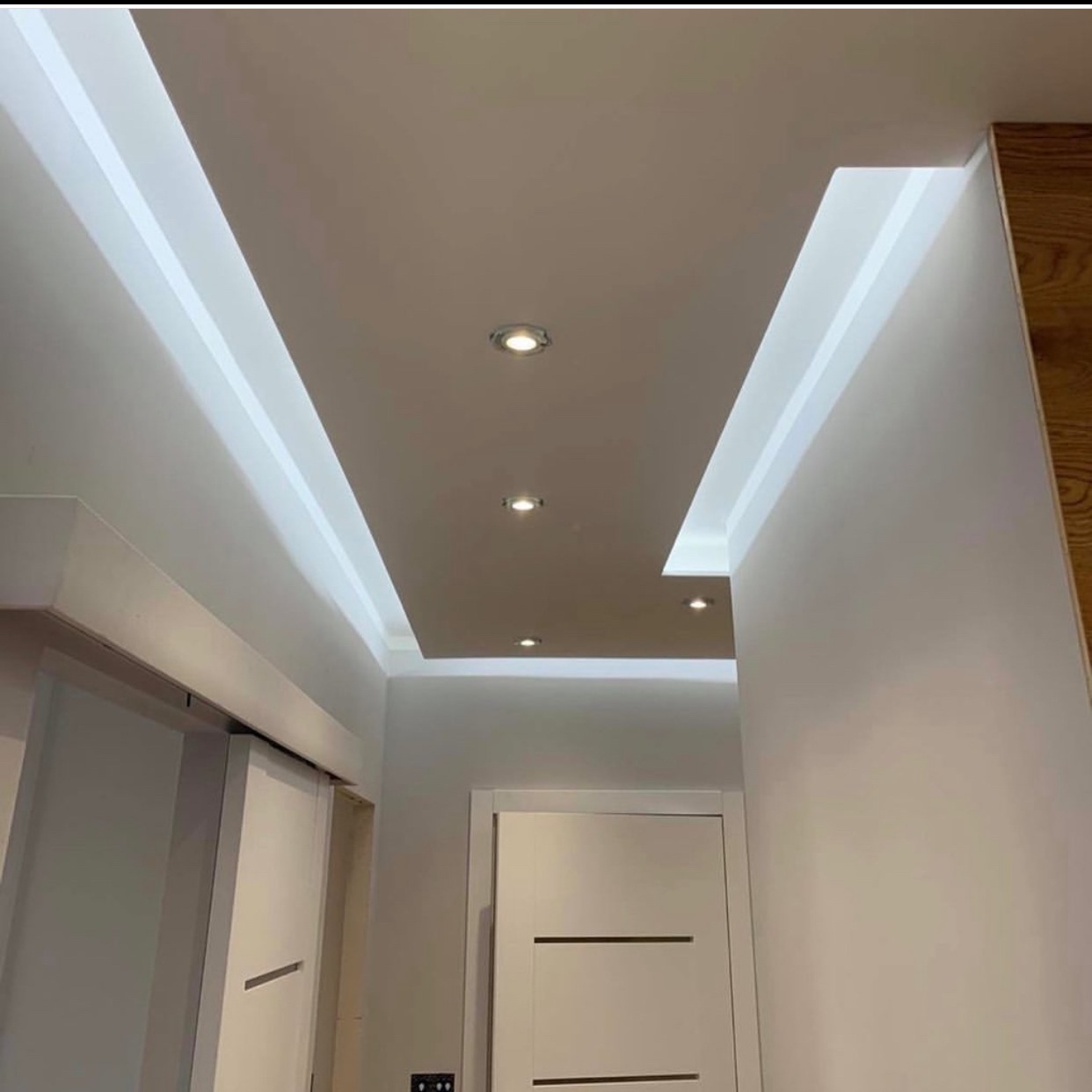 Nowoczesny sufit podwieszany z oświetleniem LED i punktowymi lampami, białe ściany, minimalistyczne drzwi wewnętrzne. Detal wykończenia korytarza.