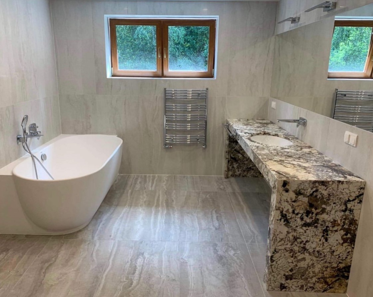 Nowoczesna łazienka z wolnostojącą wanną, marmurowym blatem umywalki i grzejnikiem drabinkowym. Jasne płytki imitujące marmur na ścianach i podłodze.