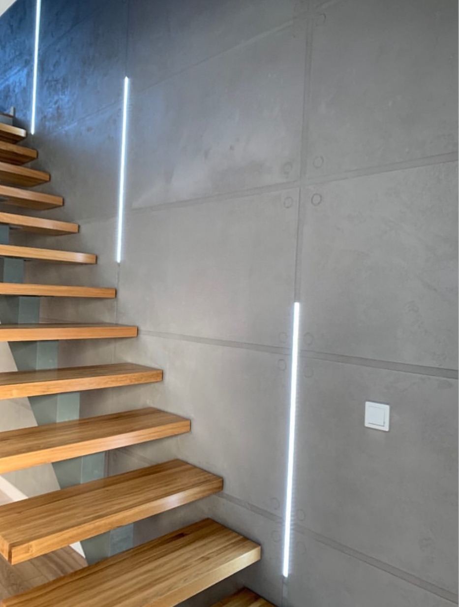 Nowoczesne schody drewniane z podświetleniem LED na szarej ścianie z betonowych paneli. Minimalistyczny design, oświetlenie liniowe, detale wykończeniowe.