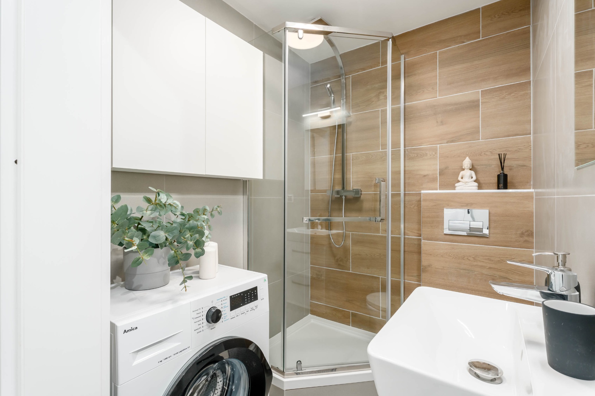 Jasna łazienka z kabiną prysznicową, pralką Amica i drewnopodobnymi płytkami. Na pralce dekoracje: roślina w doniczce i świeca. Minimalistyczny styl.