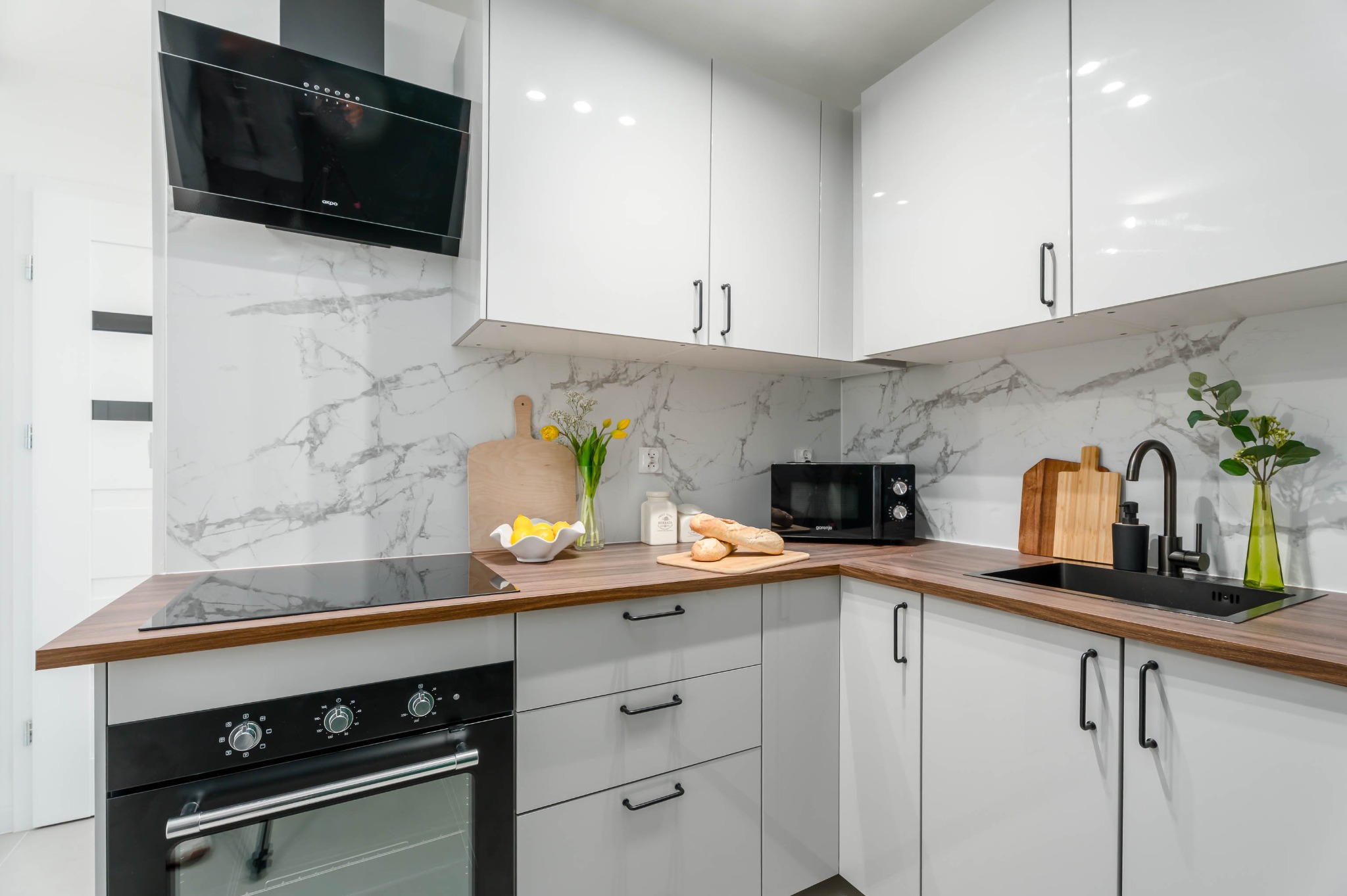 Nowoczesna kuchnia w bieli i szarości z drewnianym blatem. Marmurowe wykończenie ściany, czarne akcenty armatury i sprzętu AGD. Ustawione dekoracje i akcesoria.
