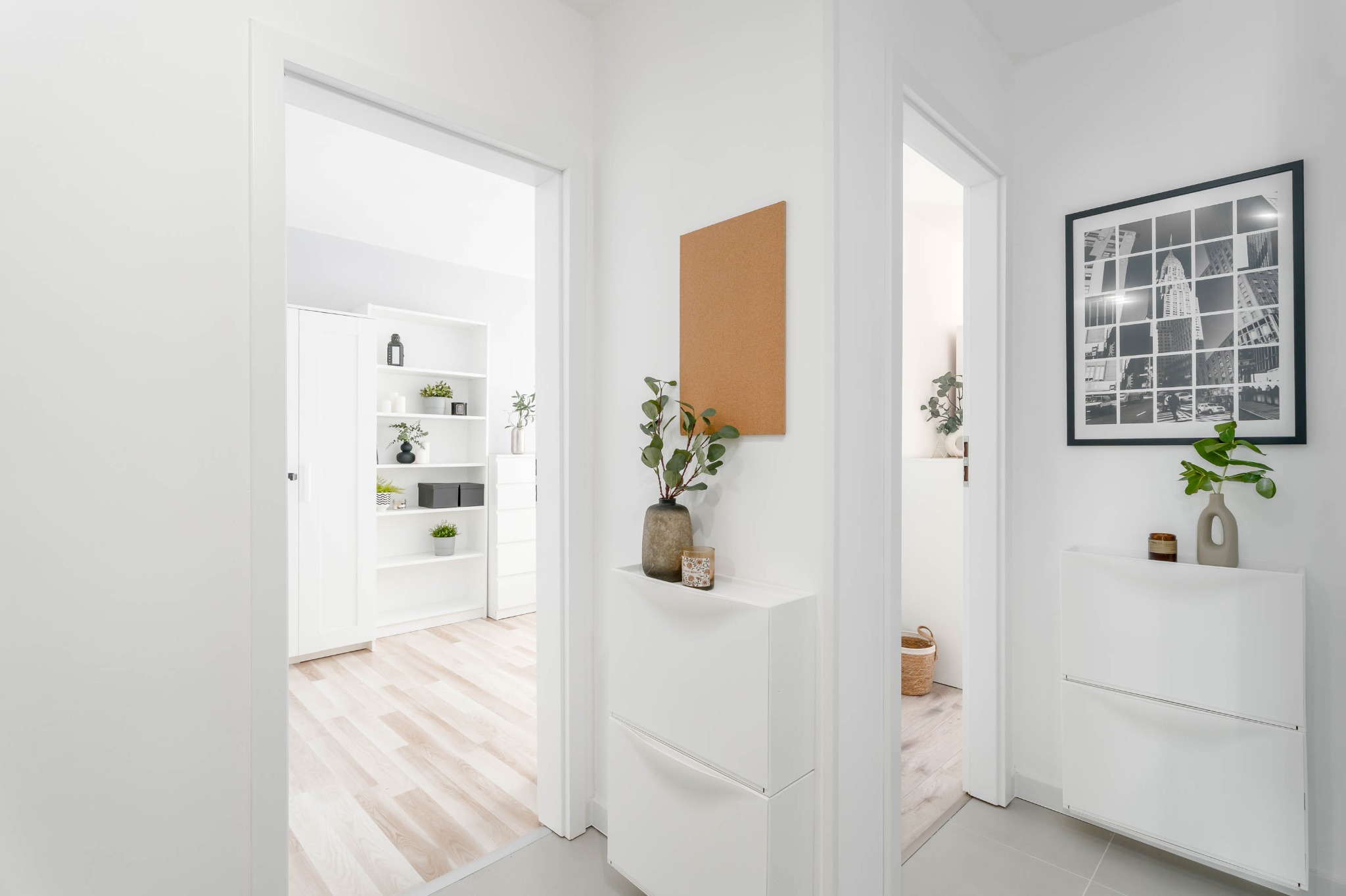 Jasne wnętrze przedpokoju z białymi meblami, roślinami w wazonach i czarno-białą fotografią w ramie. Widoczne otwarte drzwi do innych pomieszczeń.