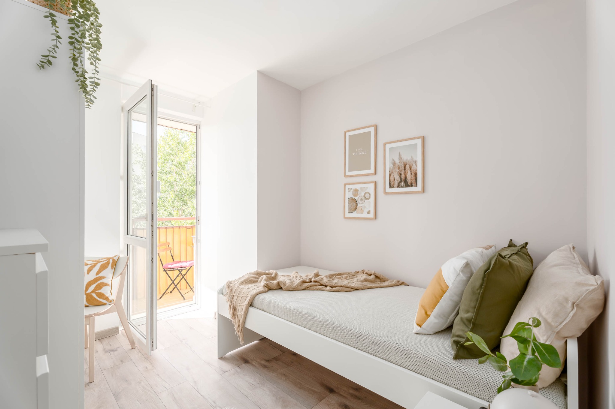 Jasny pokój z łóżkiem, otwartym balkonem i dekoracjami ściennymi w ramkach. Widoczne drewniane panele podłogowe i roślina doniczkowa. Minimalistyczny design.