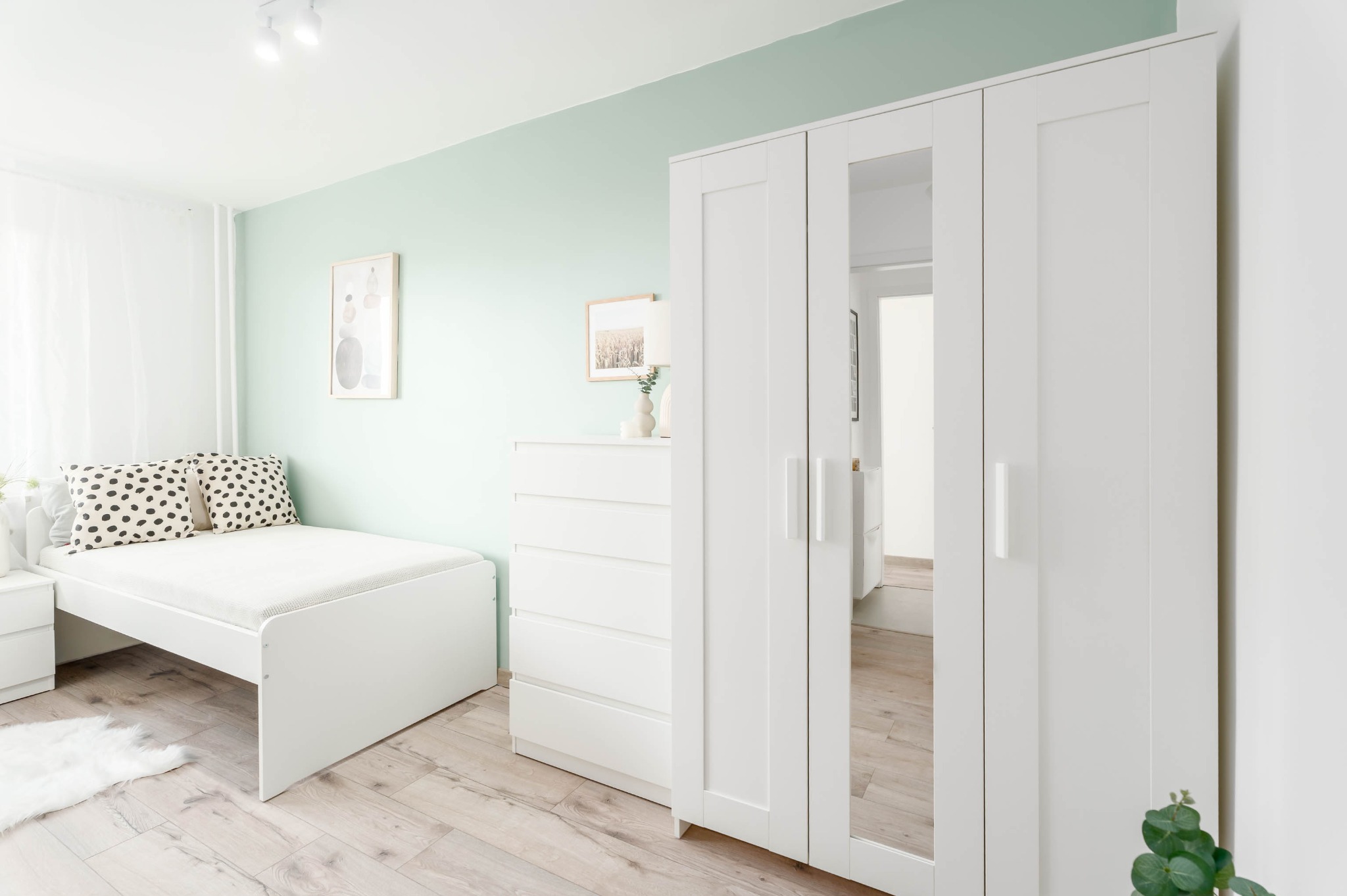 Jasne wnętrze sypialni z białymi meblami: łóżko z wzorzystymi poduszkami, komoda, szafa z lustrami. Ściana w odcieniu mięty, drewniana podłoga.
