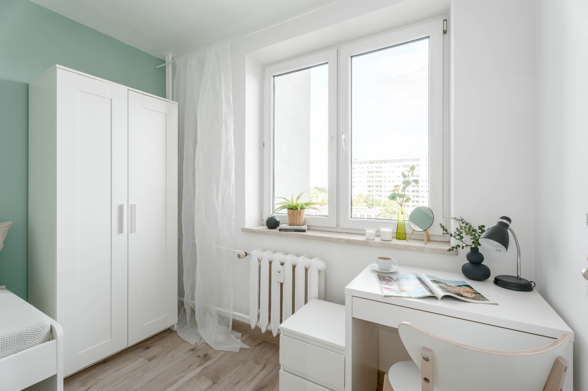 Jasne wnętrze pokoju z białymi meblami: szafa, biurko, krzesło. Okno z roślinami i dekoracjami. Drewniana podłoga i miętowa ściana dodają świeżości.