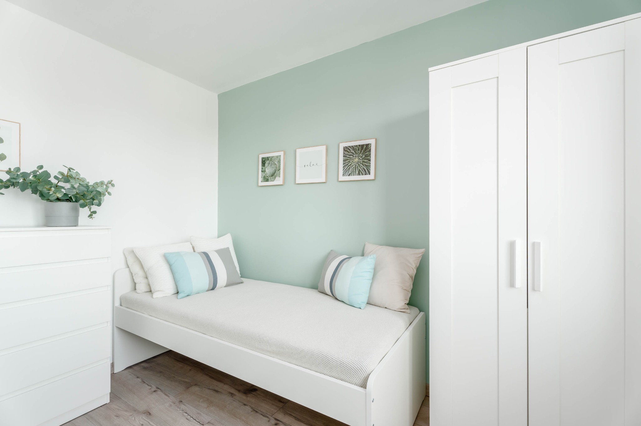 Jasny pokój z łóżkiem, komodą i szafą w bieli. Ściana w odcieniu mięty z trzema obrazkami. Drewniana podłoga. Minimalistyczny, skandynawski styl.