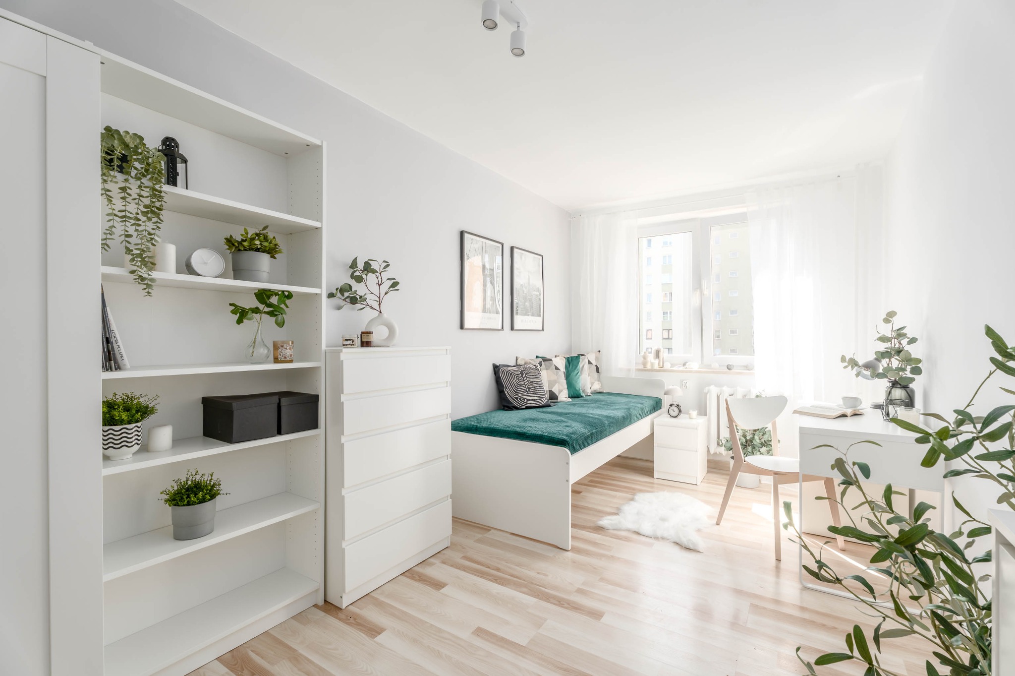 Jasne, minimalistyczne wnętrze pokoju z drewnianą podłogą, białymi meblami, roślinami doniczkowymi i dekoracjami. Widok na okno z białymi zasłonami.