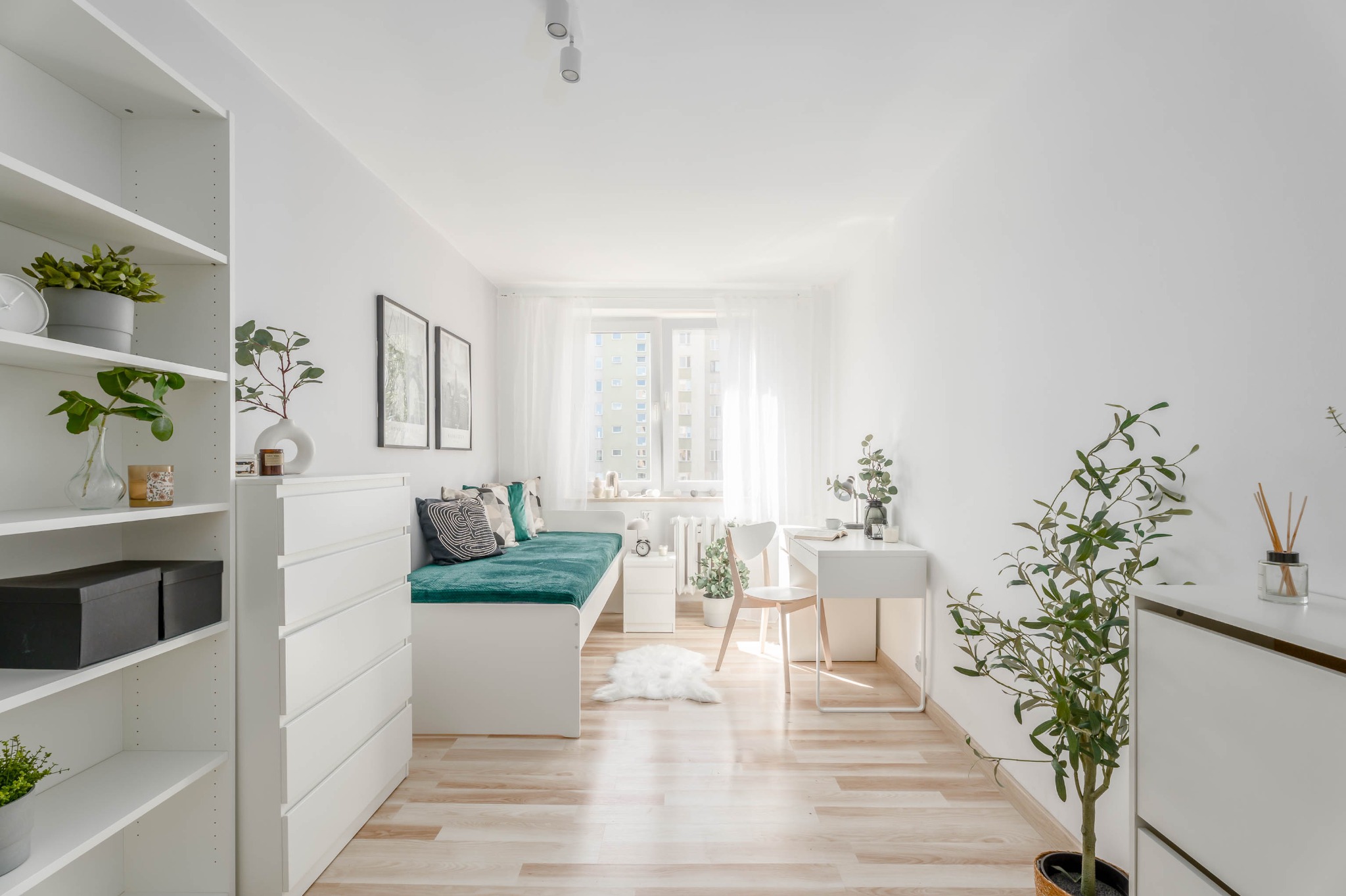 Jasne wnętrze pokoju z drewnianą podłogą, białymi meblami i zielonym akcentem na łóżku. Minimalistyczny styl, roślina doniczkowa dodaje świeżości.