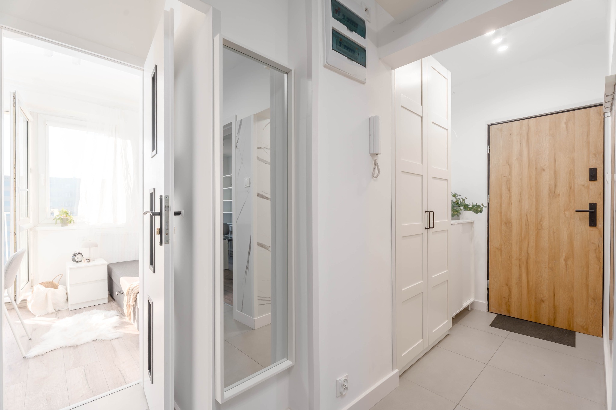 Jasne wnętrze przedpokoju z drewnianymi drzwiami wejściowymi, białymi szafami i lustrem odbijającym pokój z balkonem. Minimalistyczny design, neutralna kolorystyka.