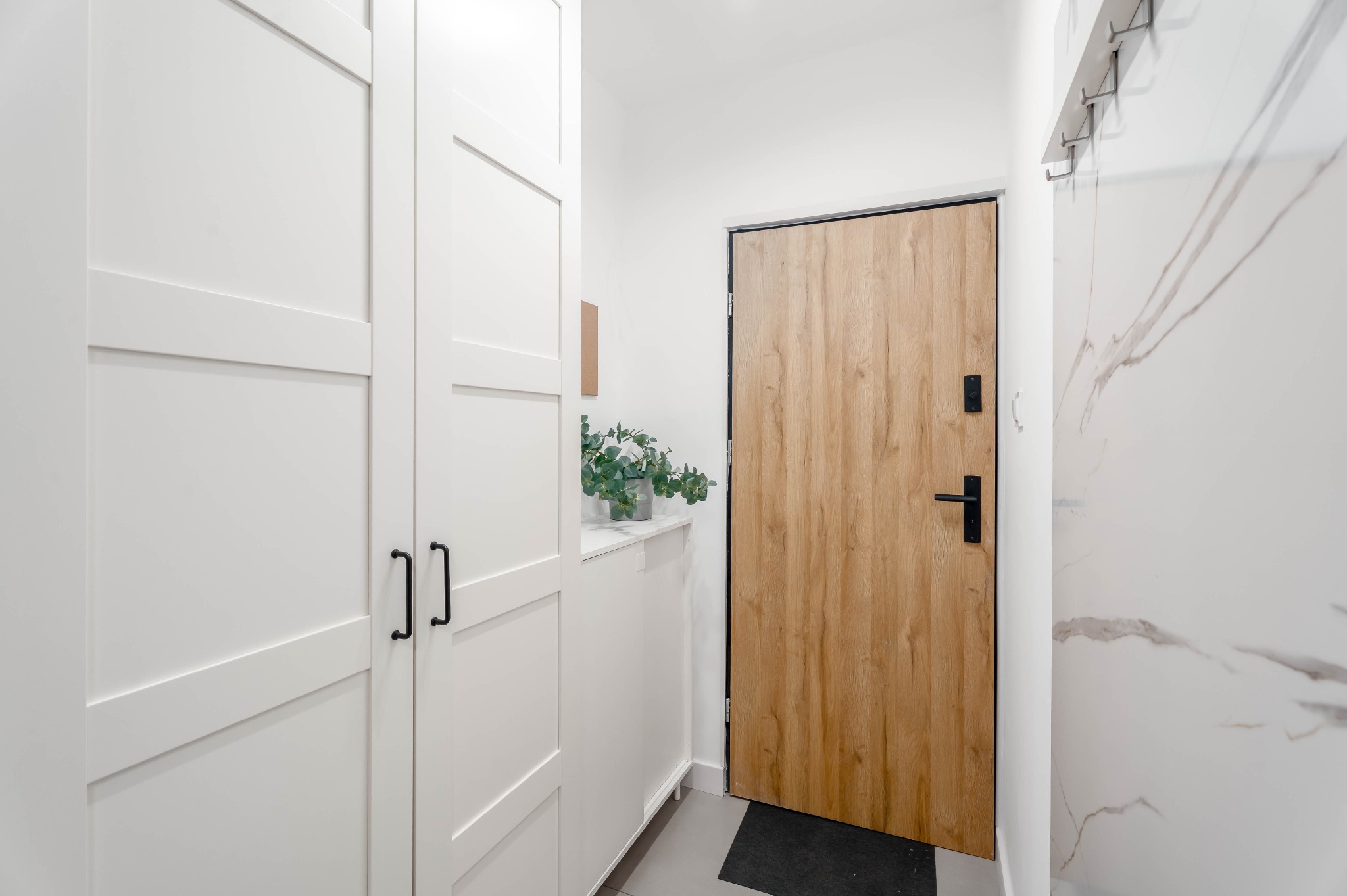 Jasne wnętrze przedpokoju z drewnianymi drzwiami i białą szafą w zabudowie. Na szafce stoi doniczka z zieloną rośliną. Ściana wykończona płytą imitującą marmur.