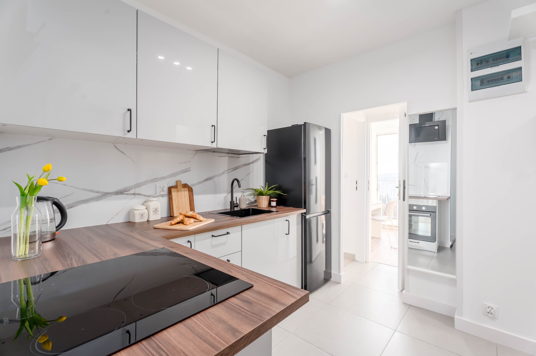 Nowoczesna, jasna kuchnia z drewnianym blatem, białymi szafkami i marmurowym backsplash. Czarna armatura i sprzęt AGD. Widok na balkon przez otwarte drzwi.