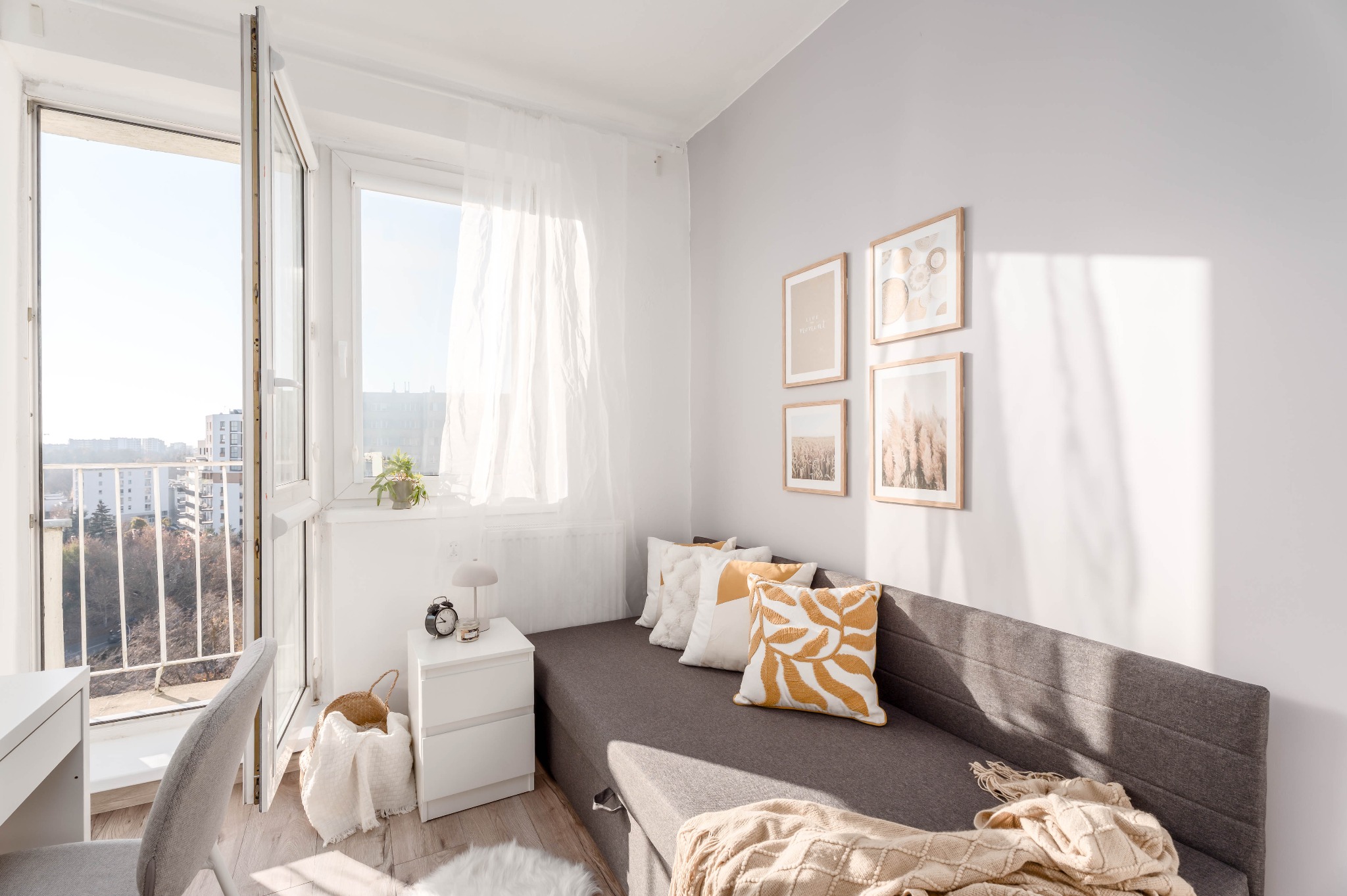Jasne wnętrze pokoju z szarą sofą, dekoracyjnymi poduszkami i kocem. Na ścianie wiszą cztery obrazy w drewnianych ramkach. Widok na balkon i miasto w tle.