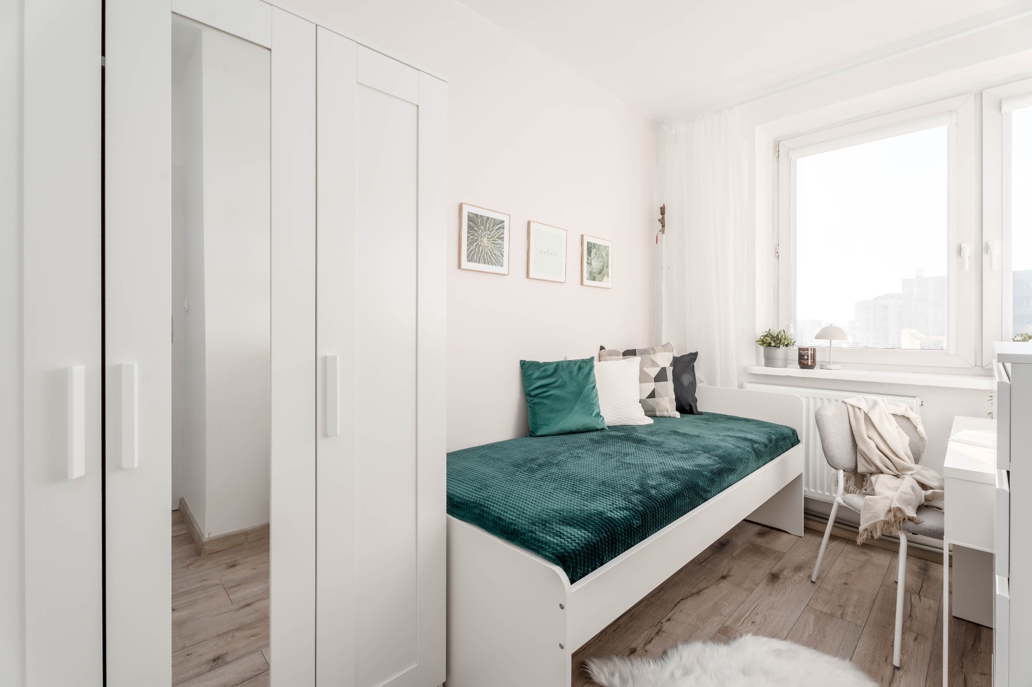 Jasne wnętrze pokoju z białymi meblami: szafa z lustrem, łóżko z zielonym pledem i poduszkami, biurko przy oknie. Drewniana podłoga i dekoracje na ścianie.