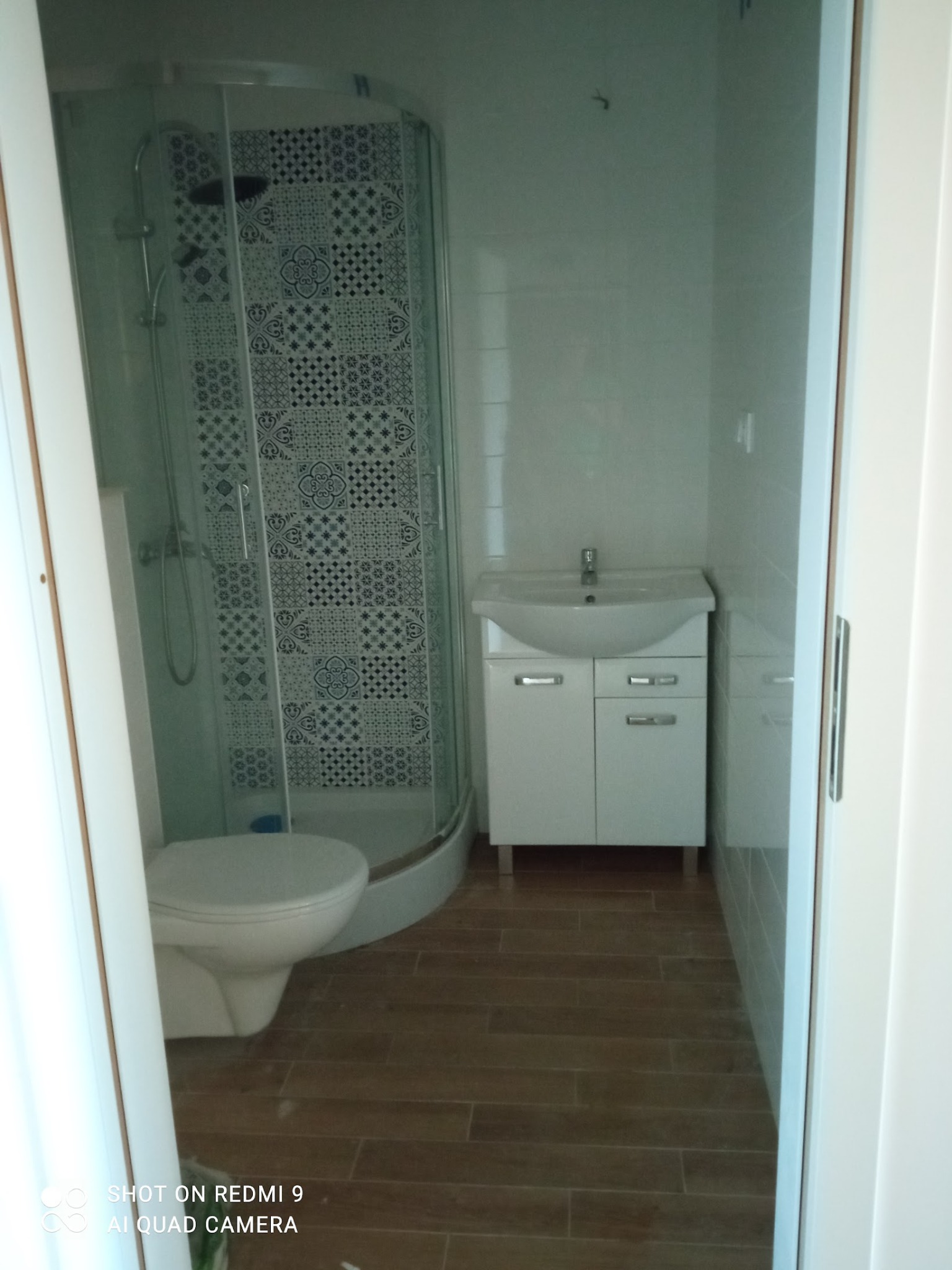 Jasna łazienka po remoncie: kabina prysznicowa z wzorzystymi płytkami, biała umywalka z szafką i toaleta. Podłoga wyłożona panelami imitującymi drewno.